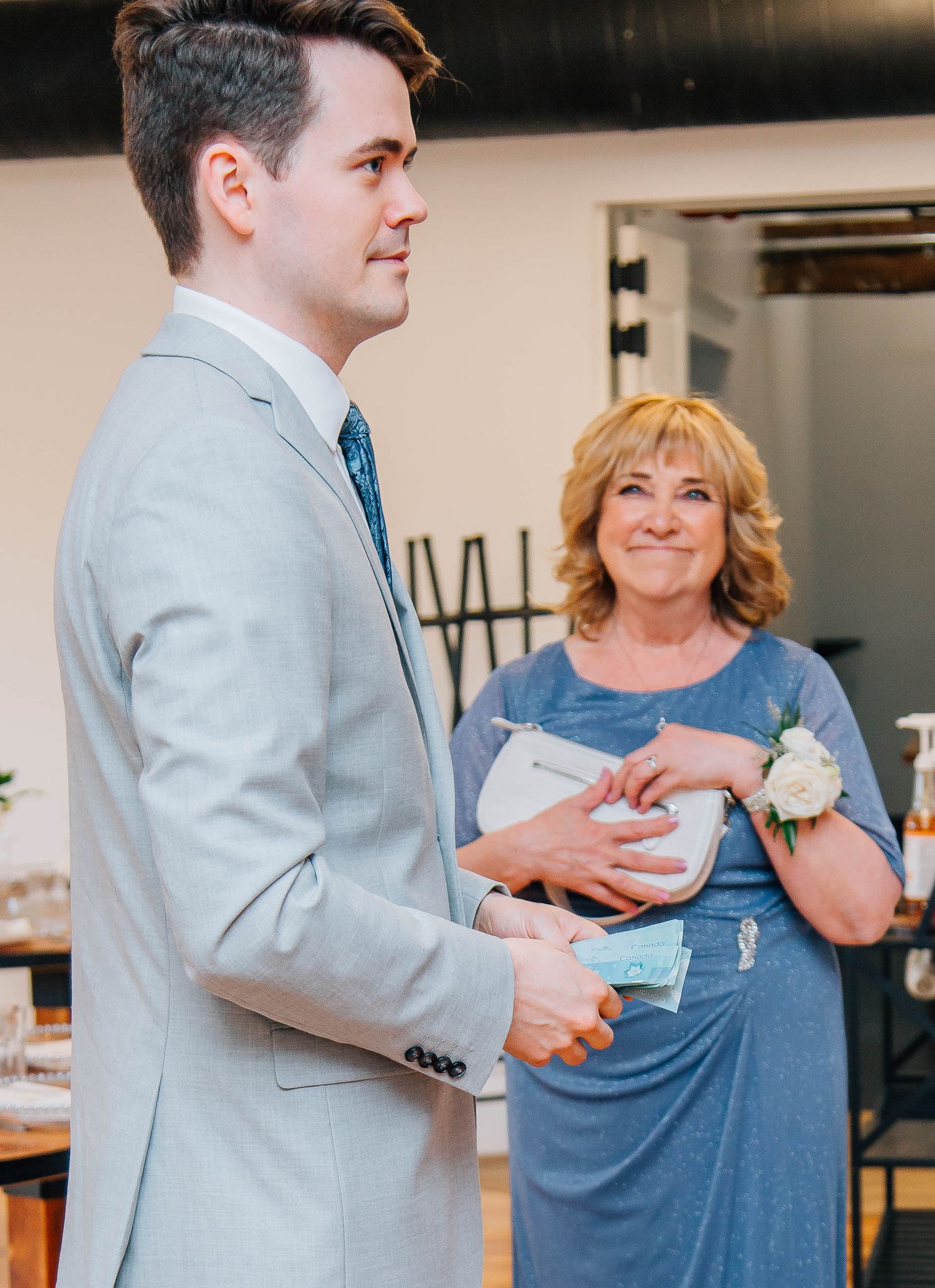 Groom in gray suit receives gift from smiling woman in blue dress at wedding venue.