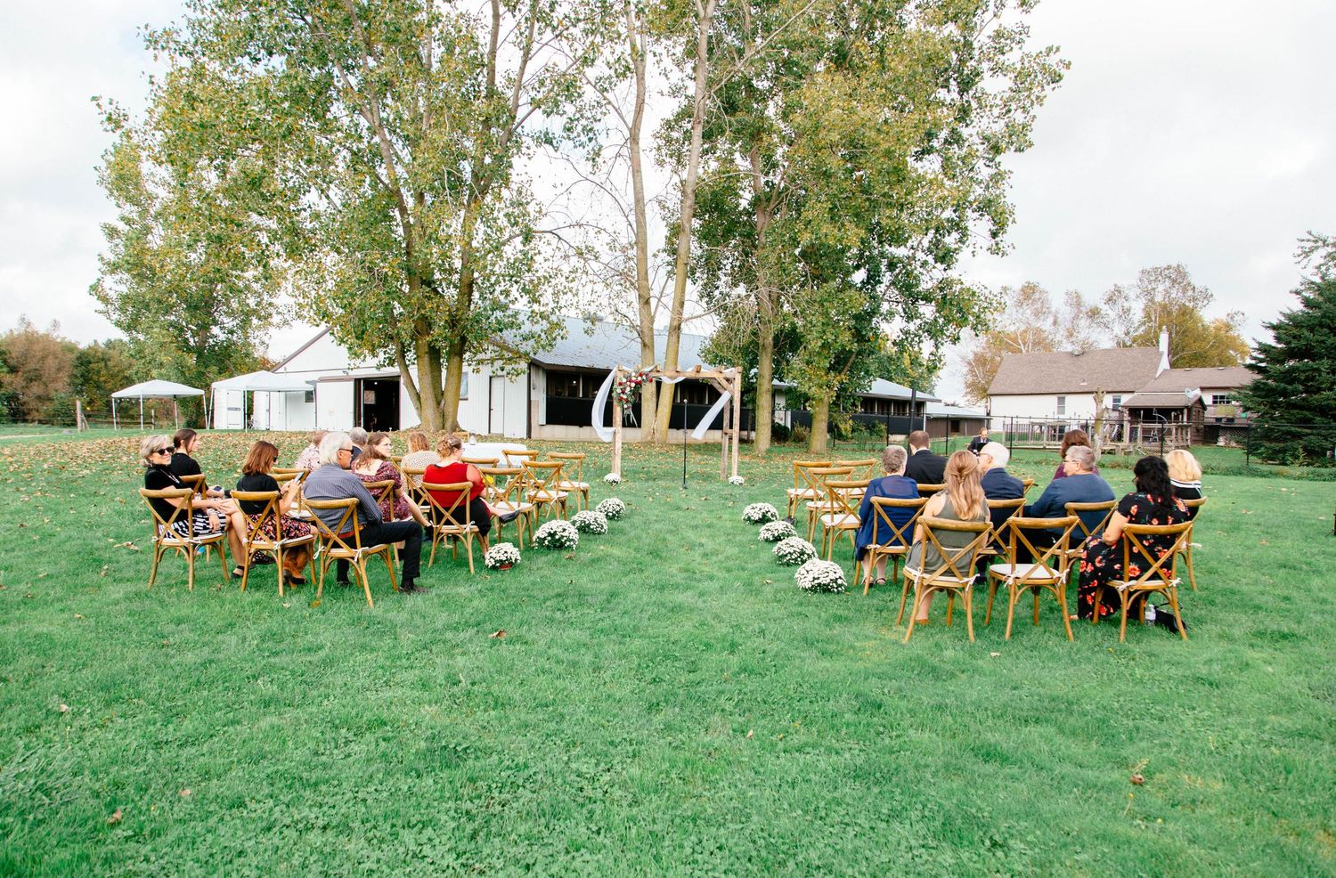 Small outdoor wedding ceremony with guests seated in wooden chairs on a green lawn surrounded by trees and farm buildings.