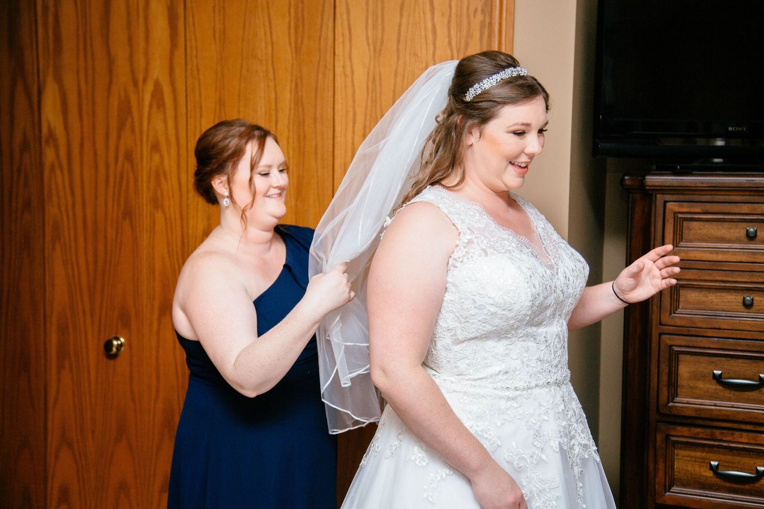 Bridesmaid in navy blue dress helping bride in lace wedding gown and veil with final preparations before ceremony.