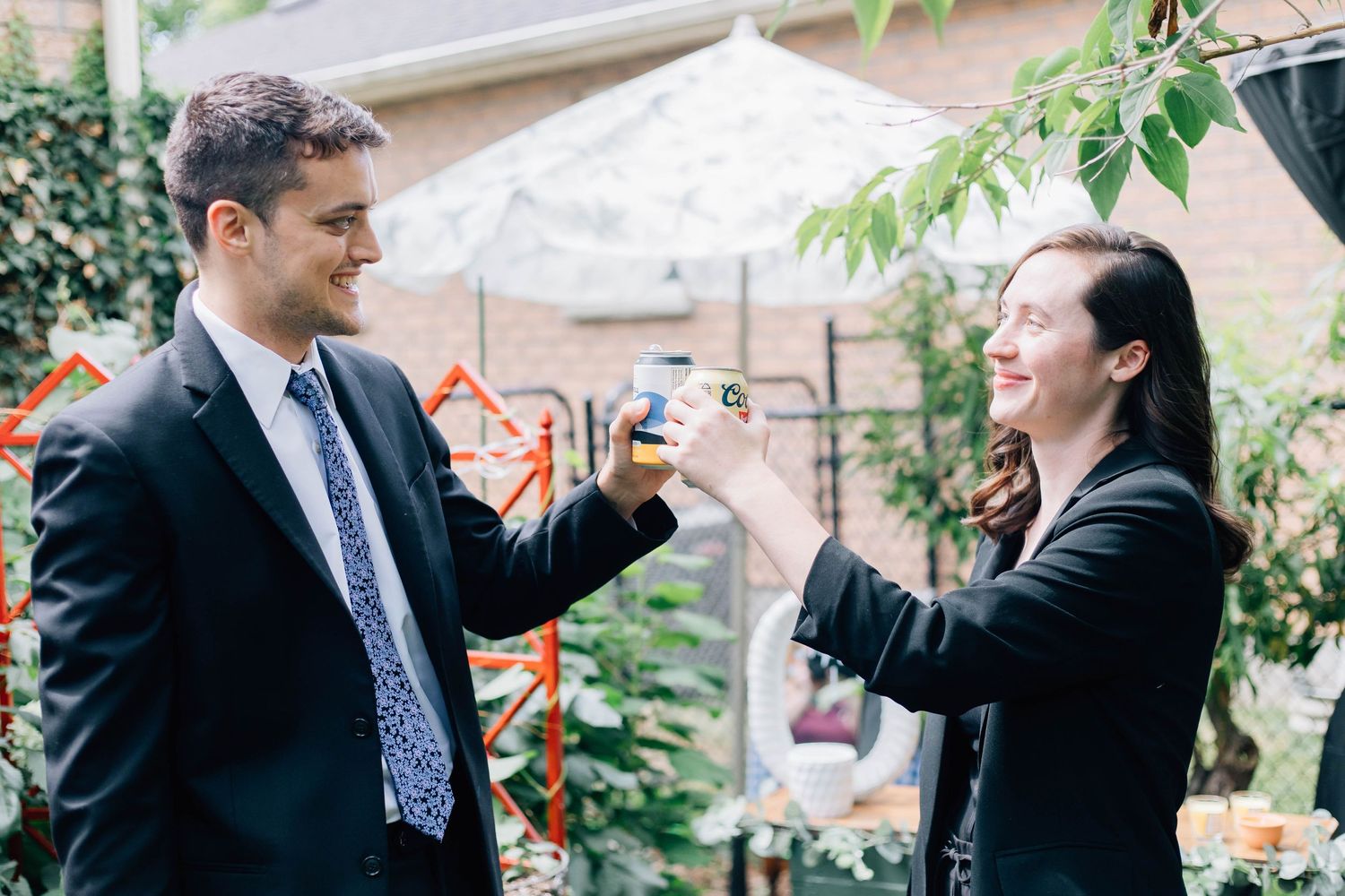 A man and woman in black suits toasting drinks outdoors at a garden wedding celebration.