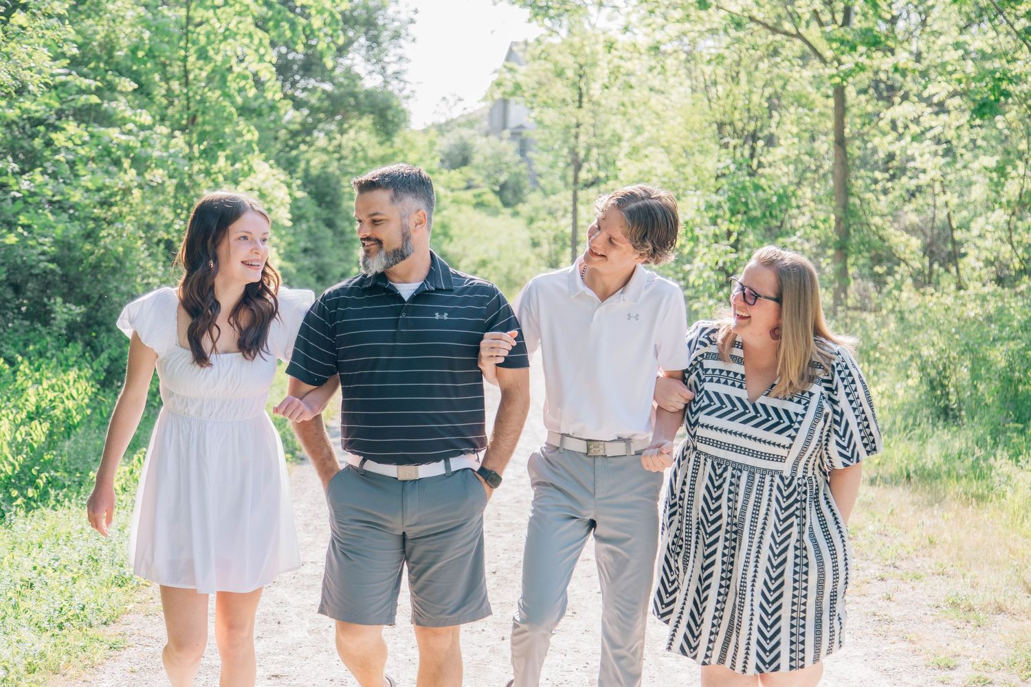 Happy family of four laughing and walking together on sunny tree-lined gravel path.