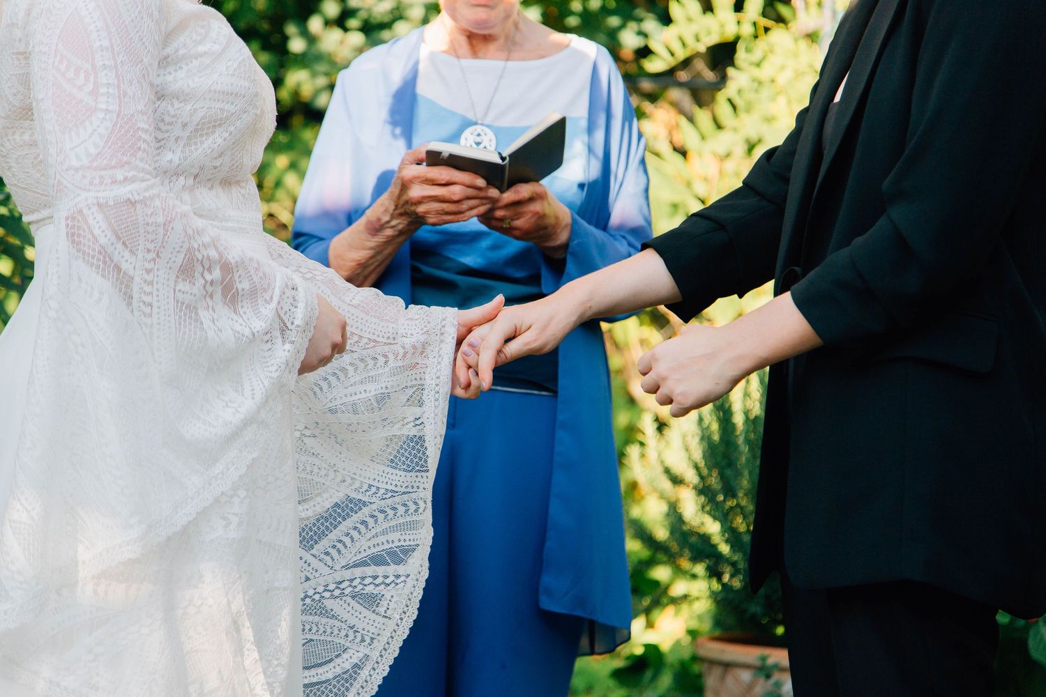 Close-up of bride and partner holding hands during ring exchange with officiant in blue dress.