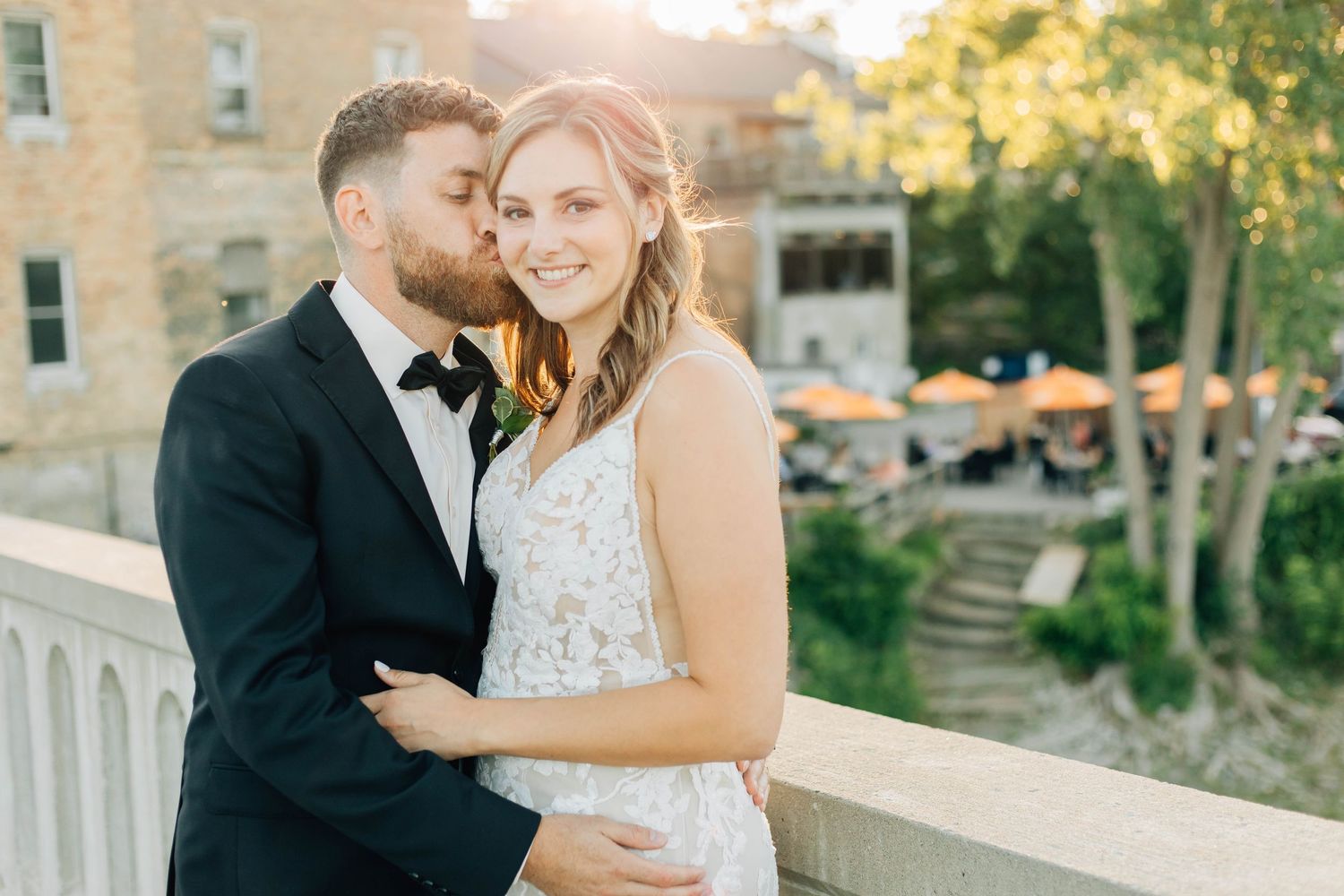 Groom tenderly kisses bride on cheek while embracing on a bridge outside Rivers Edge at the Arlington in Paris Ontario.