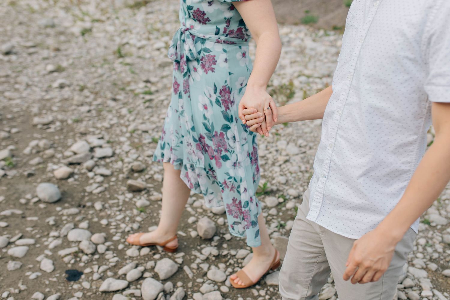 Close-up of couple holding hands while walking on rocky shore, woman in floral dress.