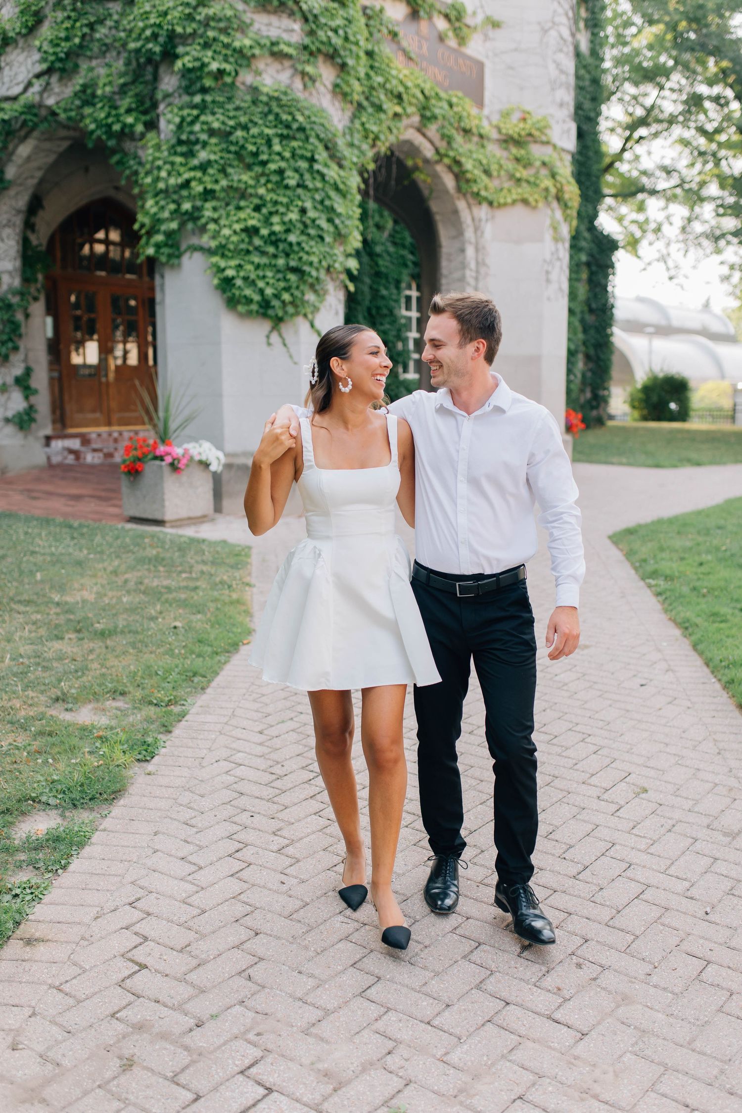 Smiling couple walks together on brick path wearing white outfits near ivy-covered stone building.