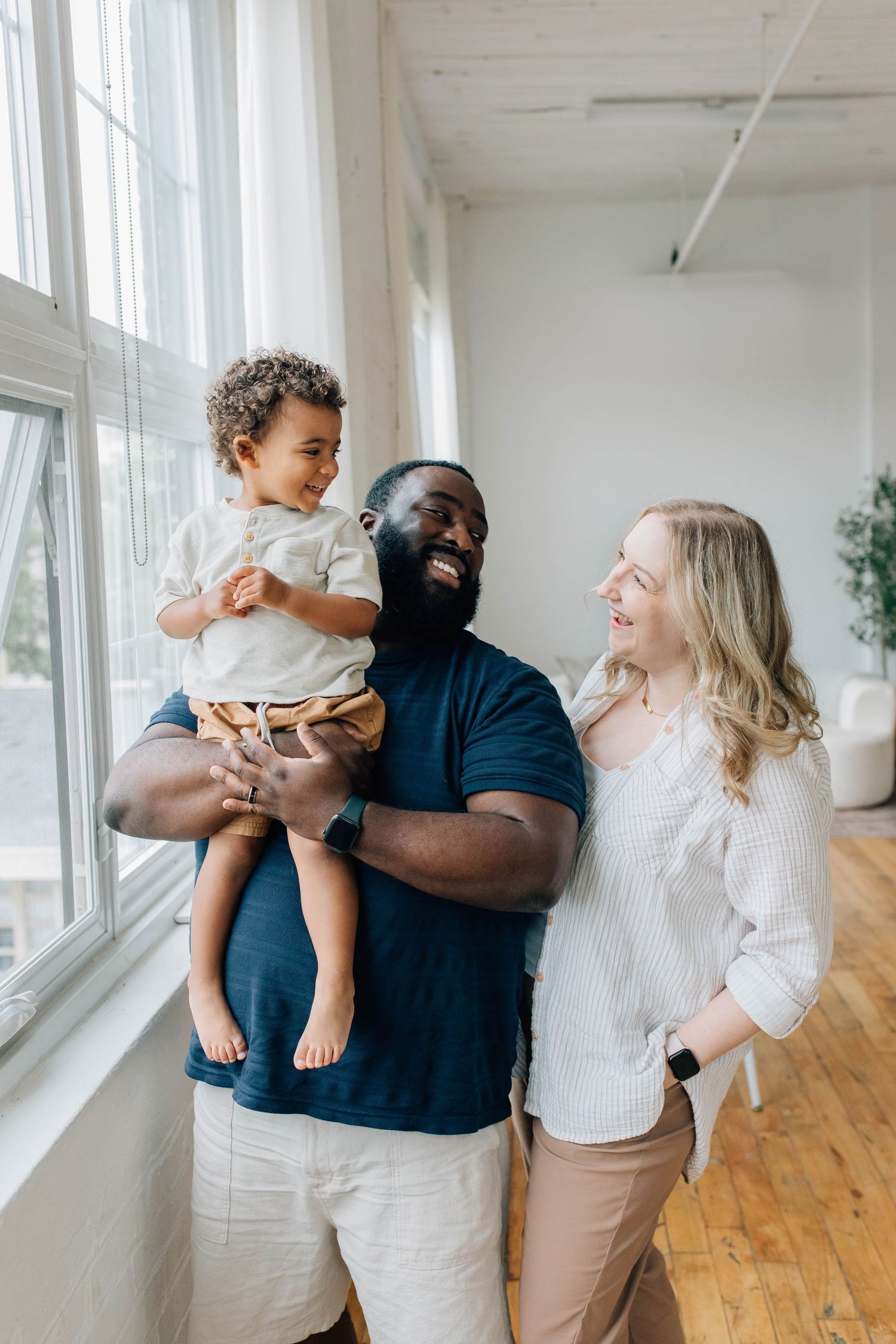 Happy interracial family with father holding toddler son while smiling with blonde mother by bright window.