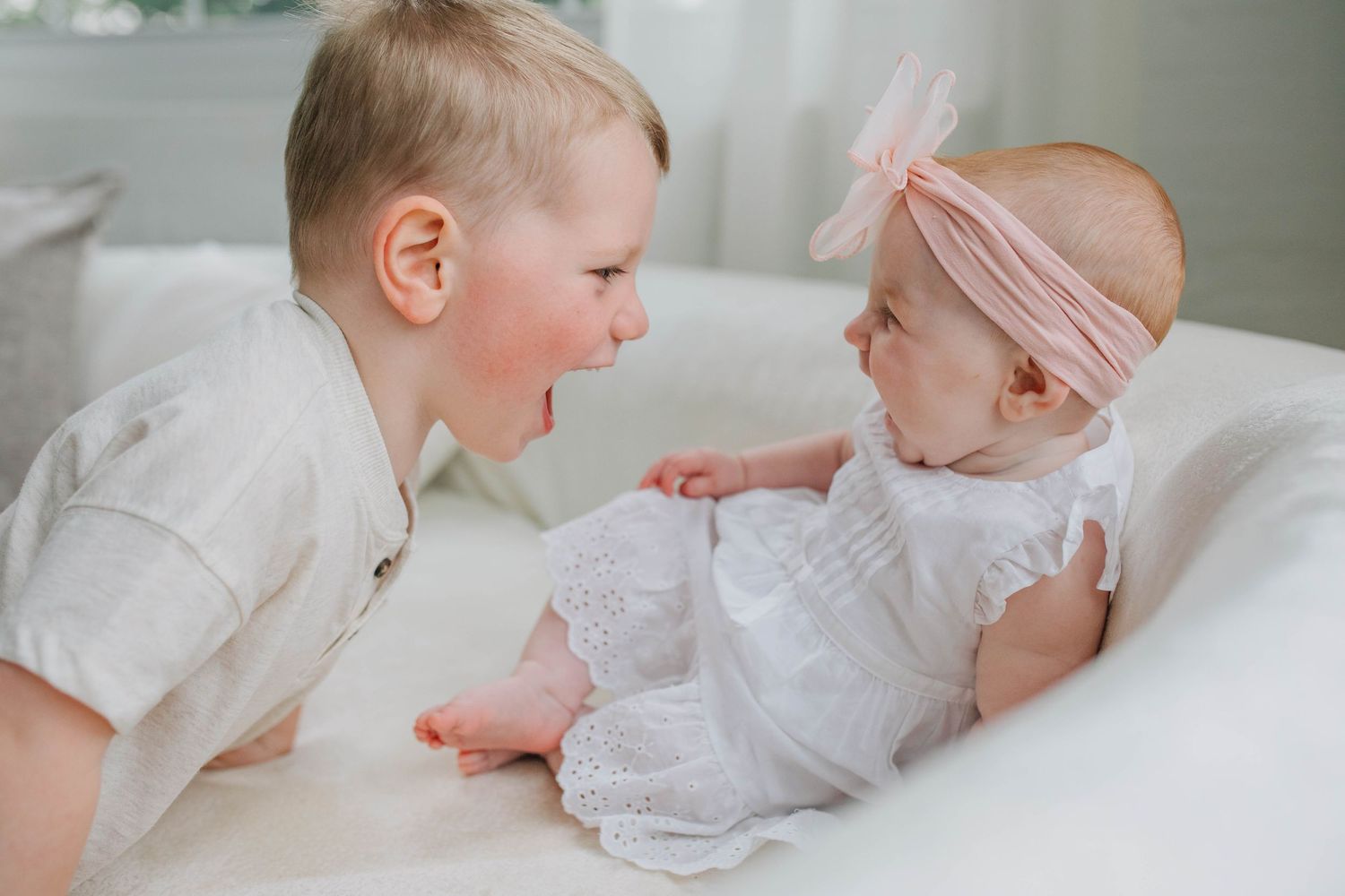 Smiling toddler boy interacting with a baby girl in a white outfit and pink headband.