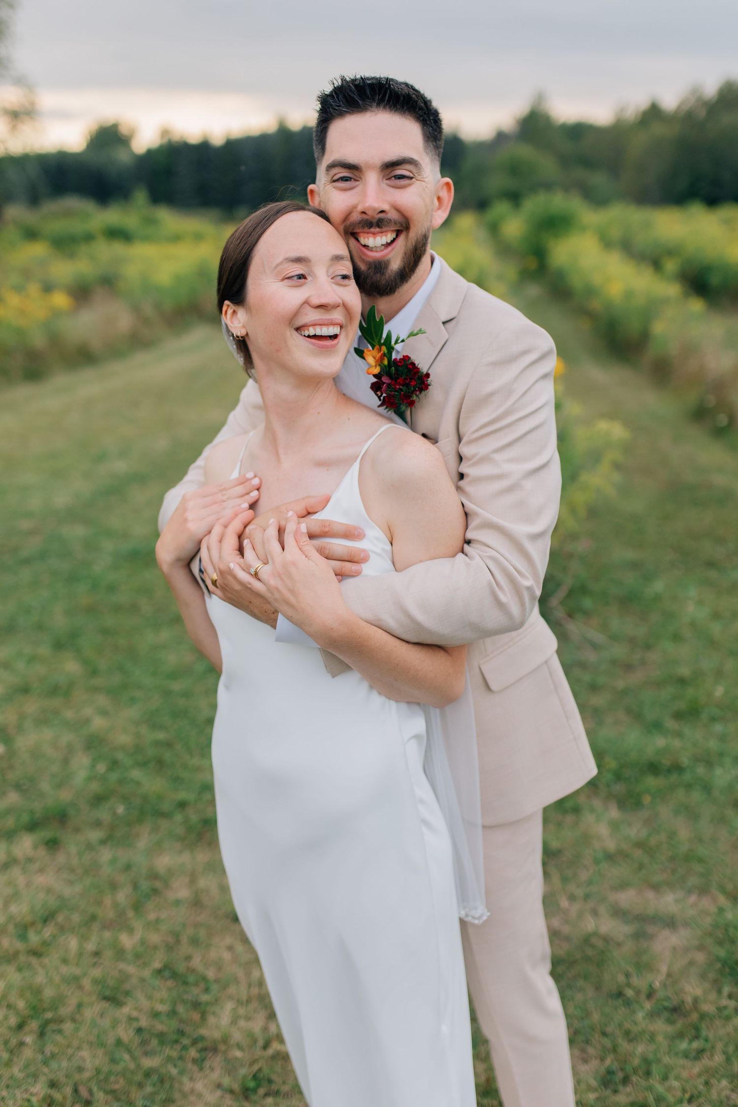 Groom in beige suit hugging laughing bride in white dress in green field at sunset.