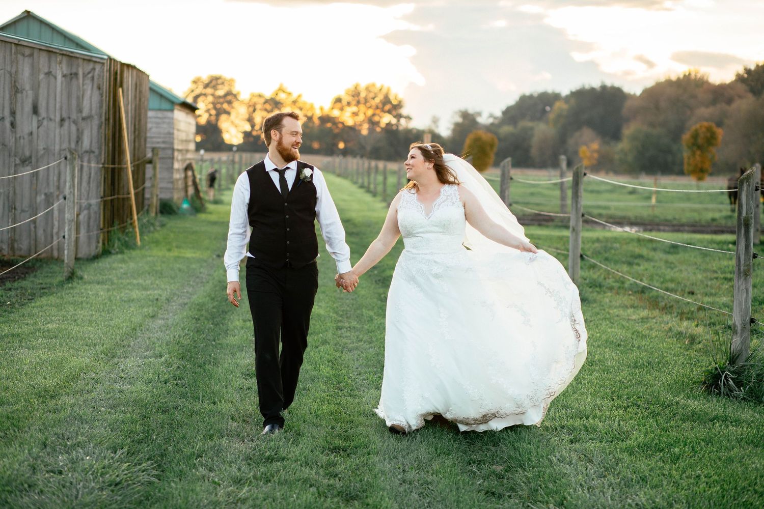 Bride and groom holding hands walking through a lush green farm field at golden hour.
