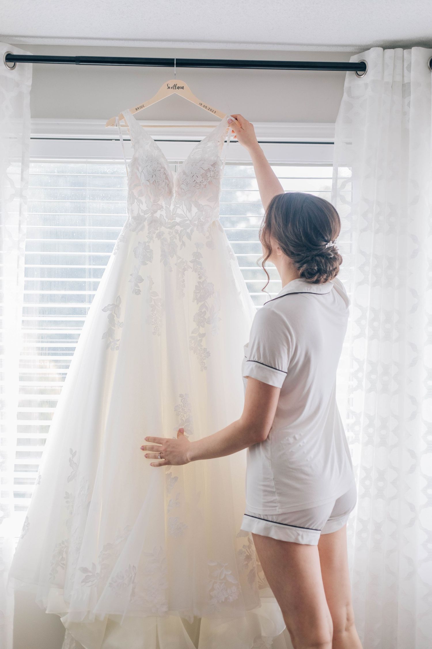 Bride in pajamas reaching up to touch her lace wedding gown hanging by window.