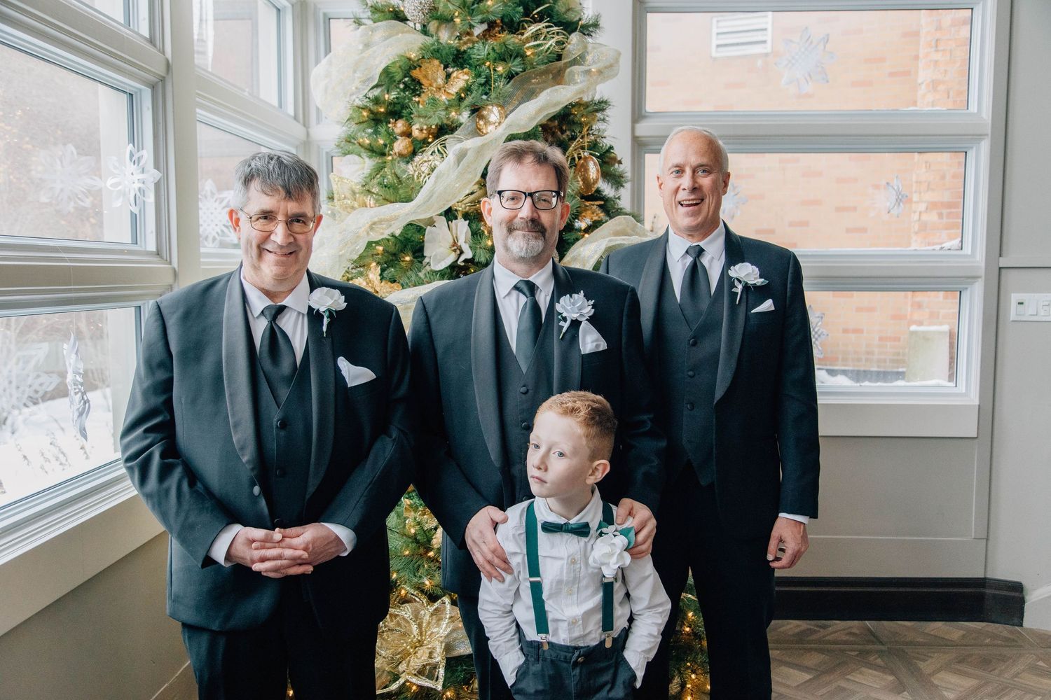 Three groomsmen in dark suits with boutonnieres pose with young ring bearer by Christmas tree at winter wedding.