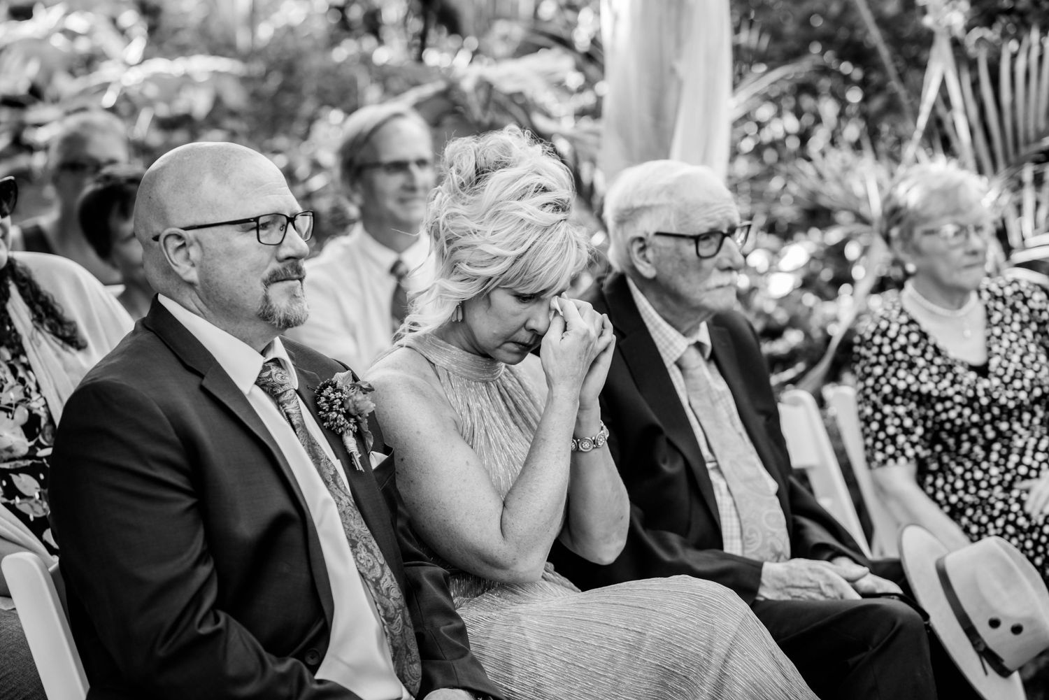 Emotional wedding guest wipes tears during ceremony seated between two men in black and white photo.