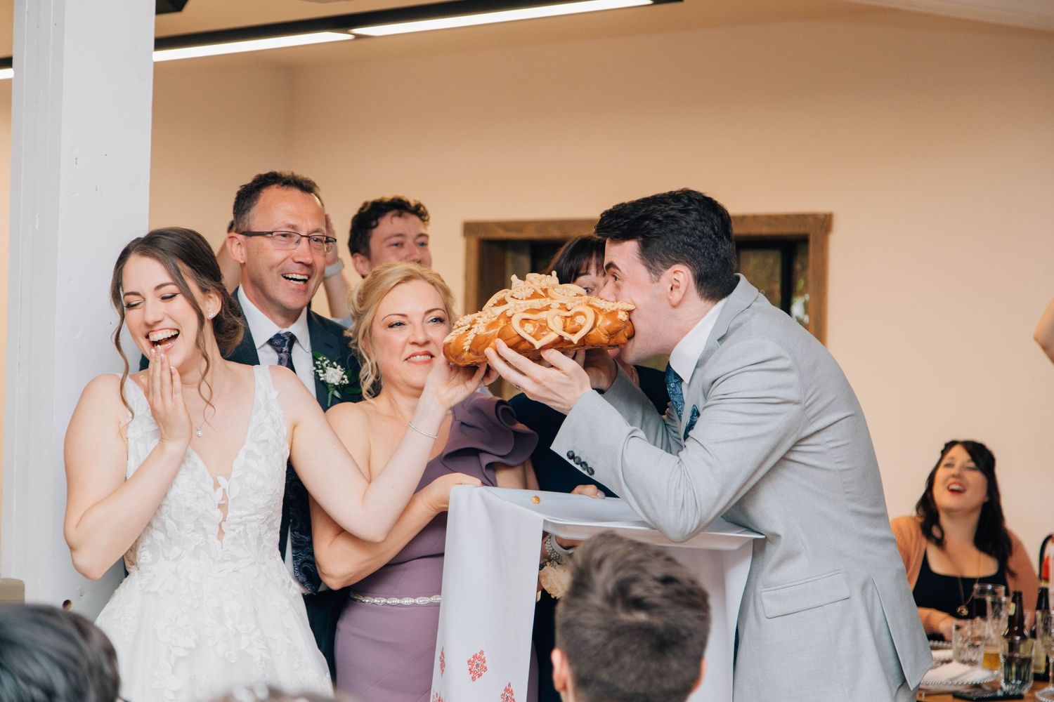 Wedding guests laughing as groom bites into a braided bread loaf at reception.