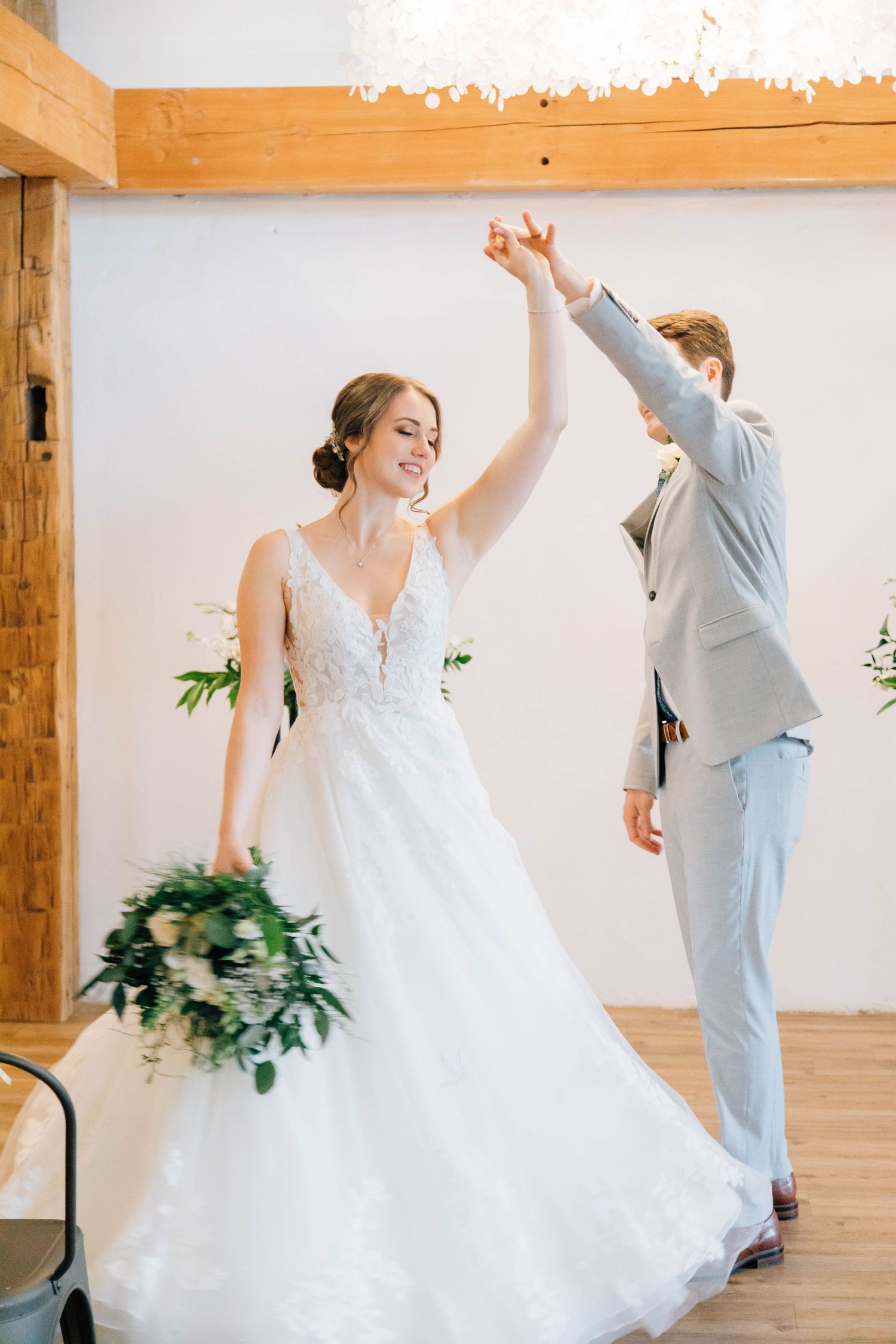 Joyful bride in lace ballgown twirling with groom in gray suit during indoor wedding dance.