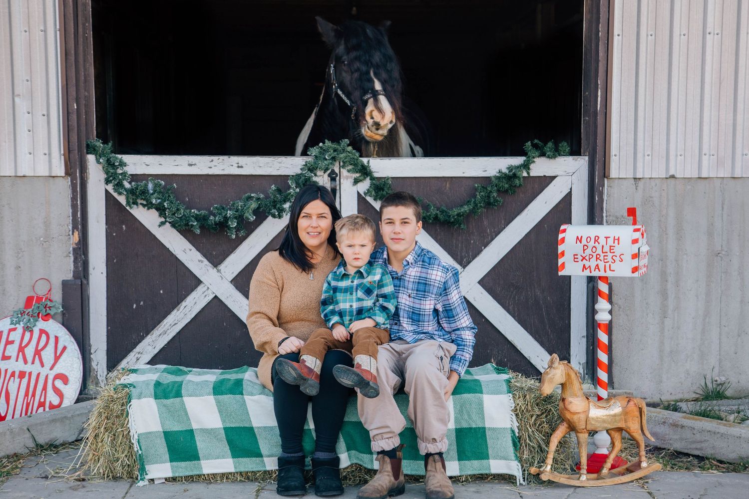Family of three sitting on hay bale in front of decorated barn with horse peeking out.