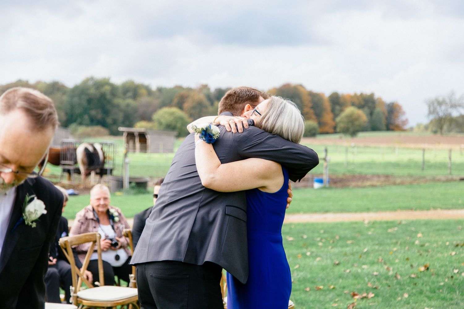 Groom in gray suit shares an emotional hug with a woman in a blue dress at an outdoor wedding ceremony.