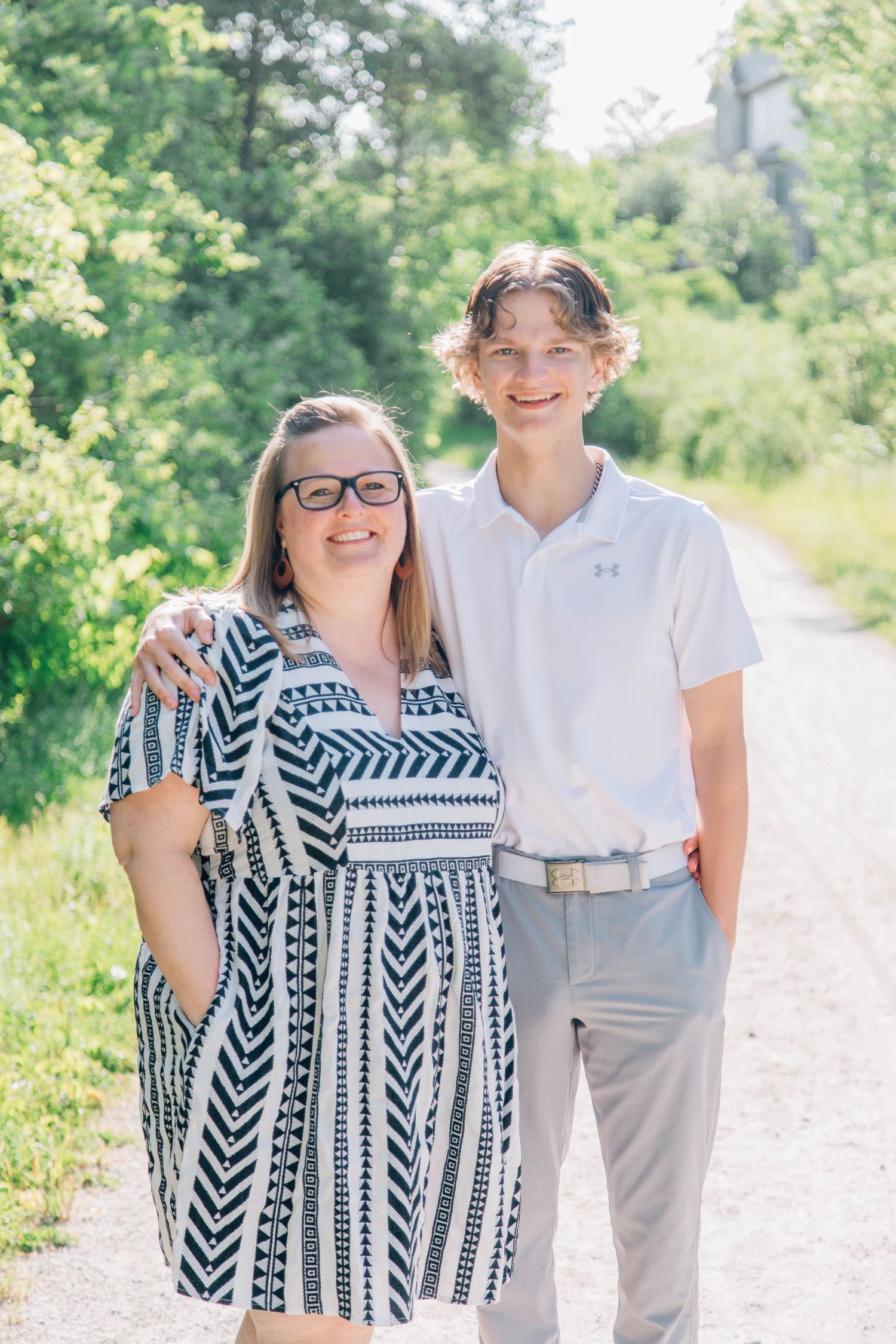 Smiling mother and teenage son posing together on sunny gravel path surrounded by greenery.