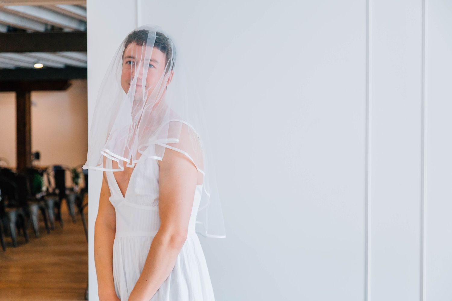 Bride in white wedding dress and veil poses elegantly against white wall at venue.