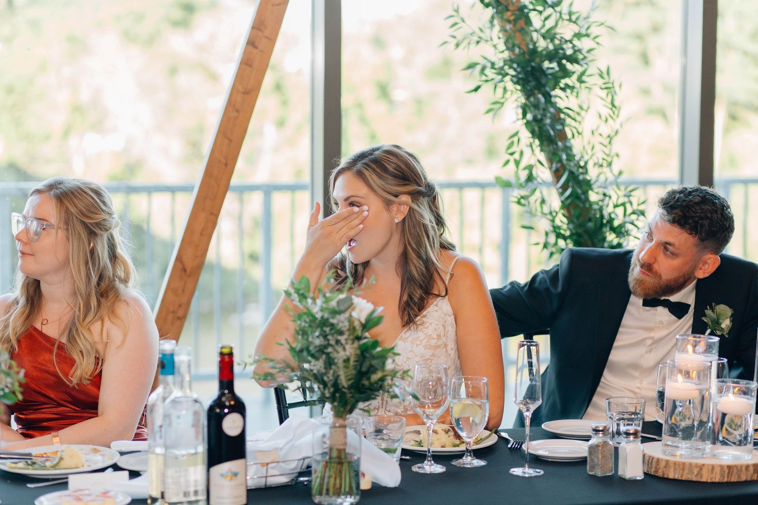 Bride laughs joyfully at head table while groom leans in during lively wedding reception with greenery decor.