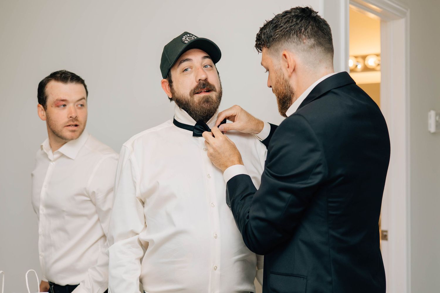 Groomsman adjusting bow tie of bearded groom wearing cap before wedding ceremony.