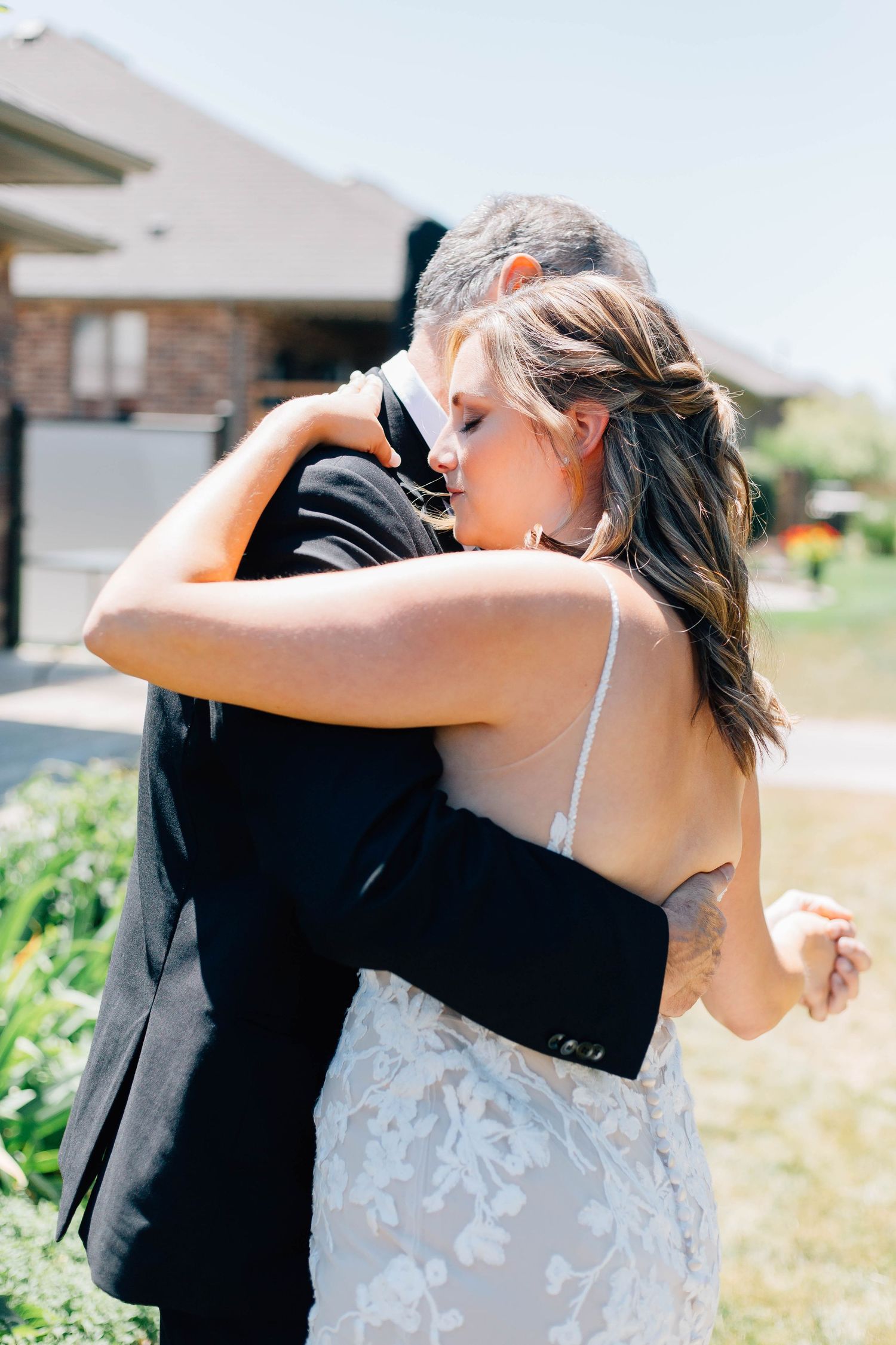 Bride in a backless lace gown shares a tender embrace with her father in a sunlit outdoor setting.
