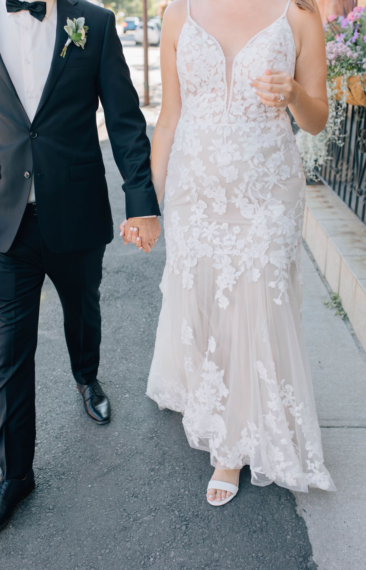 Close-up of bride in floral applique lace wedding gown holding hands with groom in black tuxedo.