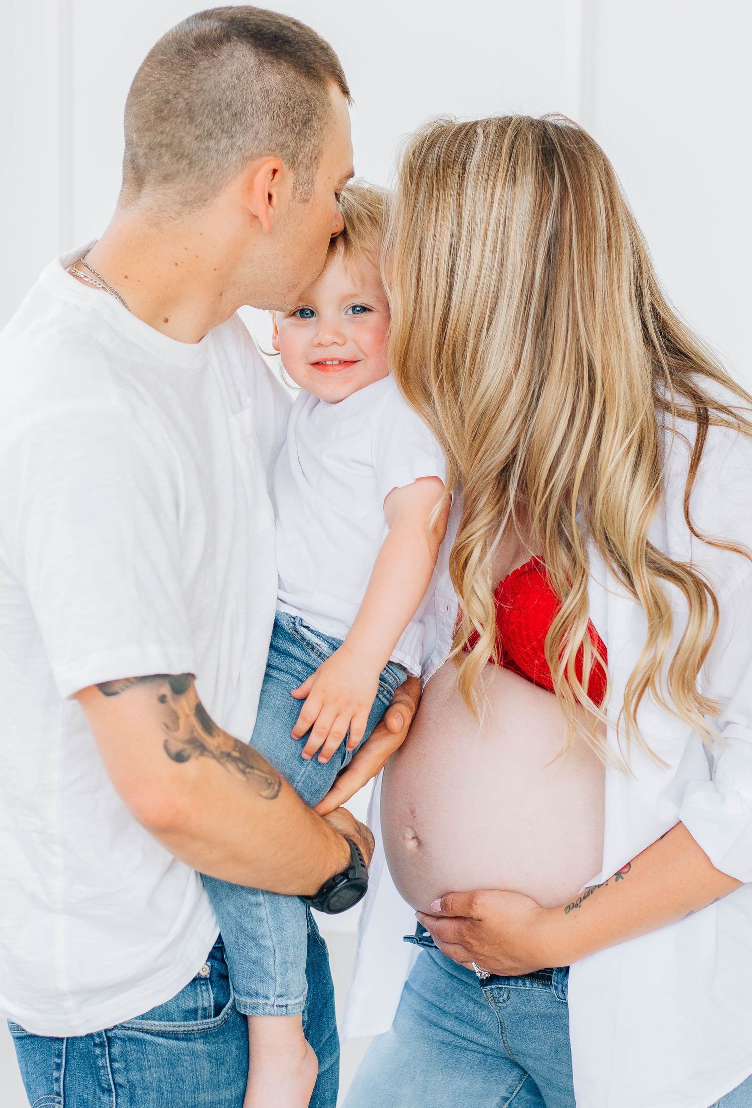 Tattooed dad kissing toddler while pregnant mom in red bralette cradles baby bump in bright white studio.