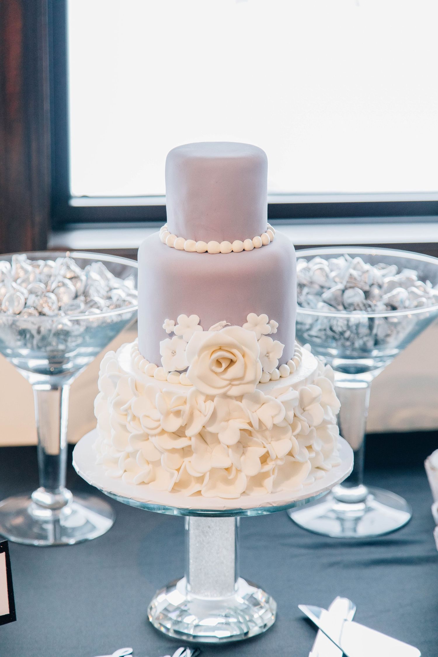 Elegant three-tier white and lavender wedding cake with ruffled flowers on a glass pedestal cake stand.