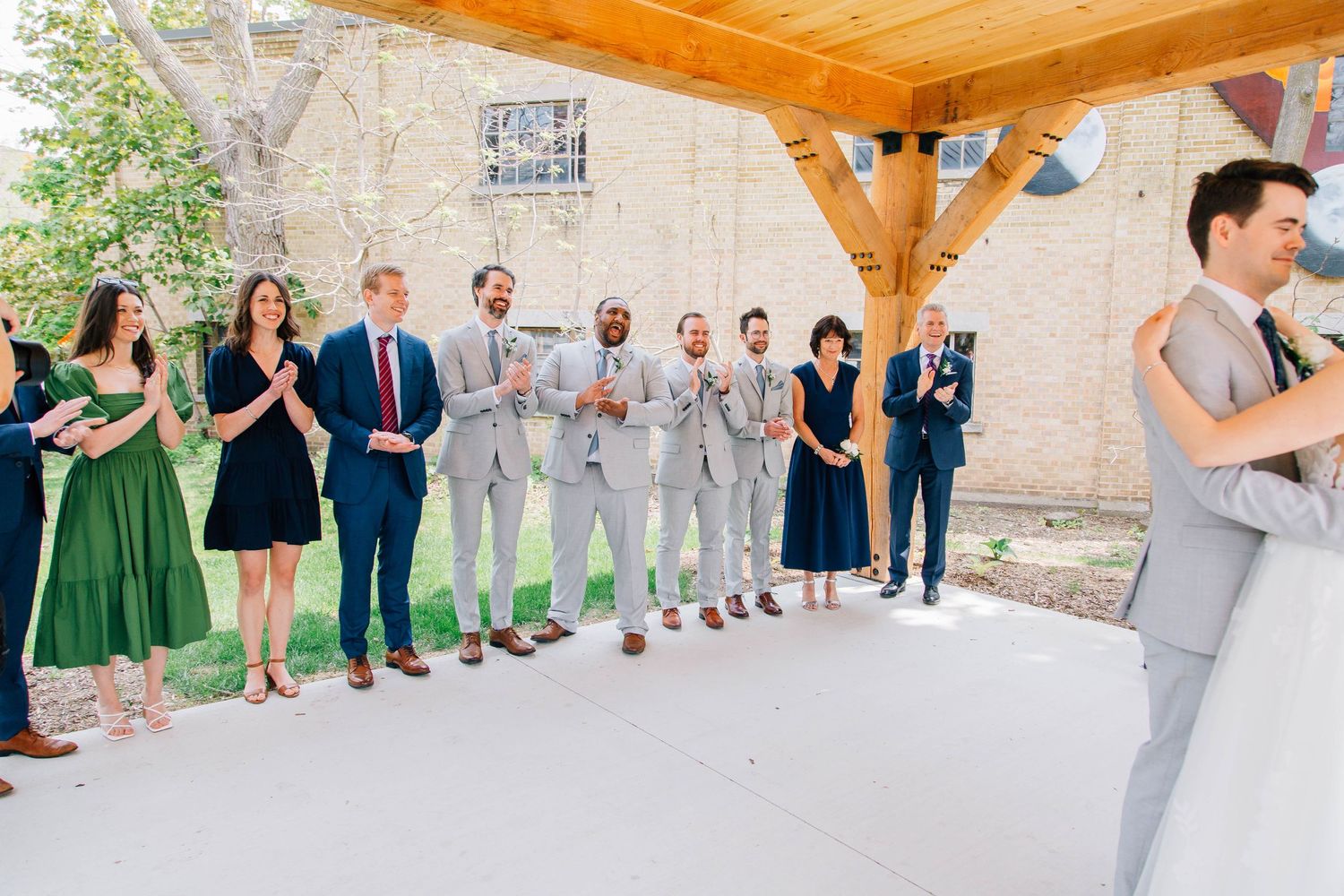 Wedding party guests applaud and celebrate under wooden pavilion during outdoor wedding ceremony.