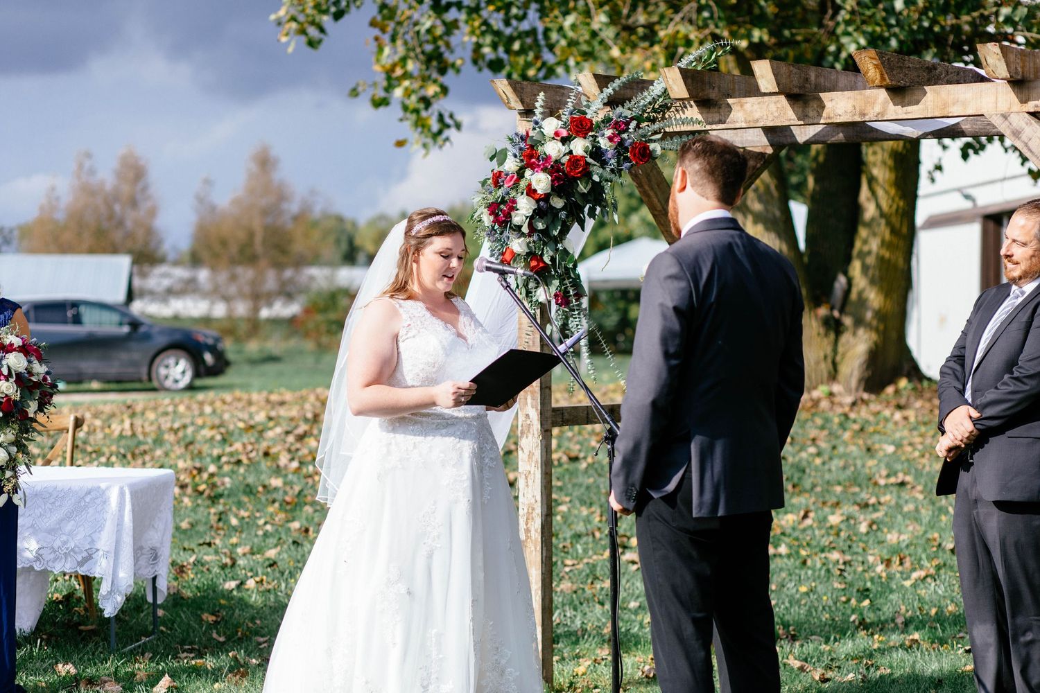 Bride reads vows from a book at rustic outdoor wedding altar adorned with red flowers.