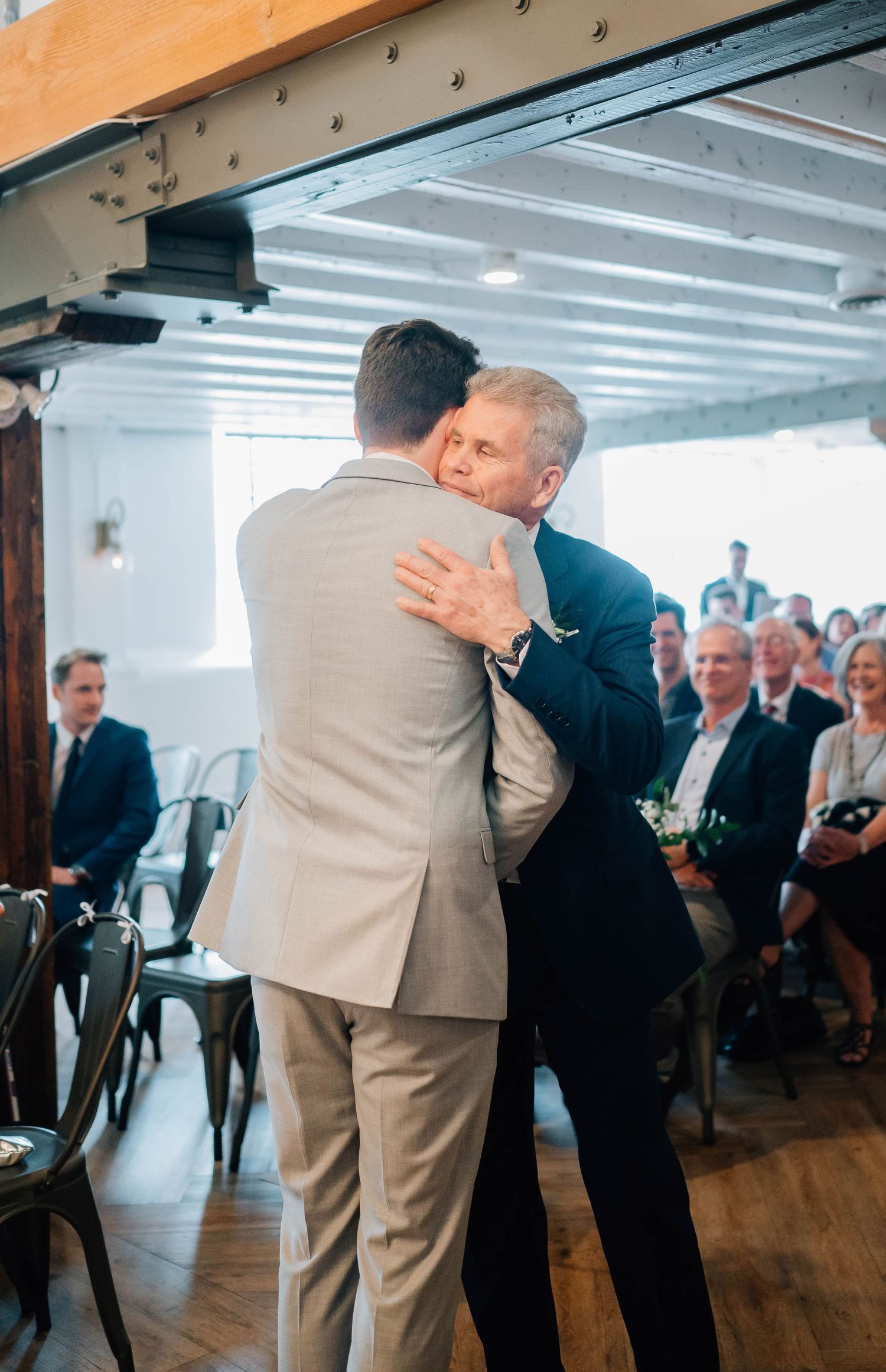 Groom embracing older man at indoor wedding ceremony with seated guests.