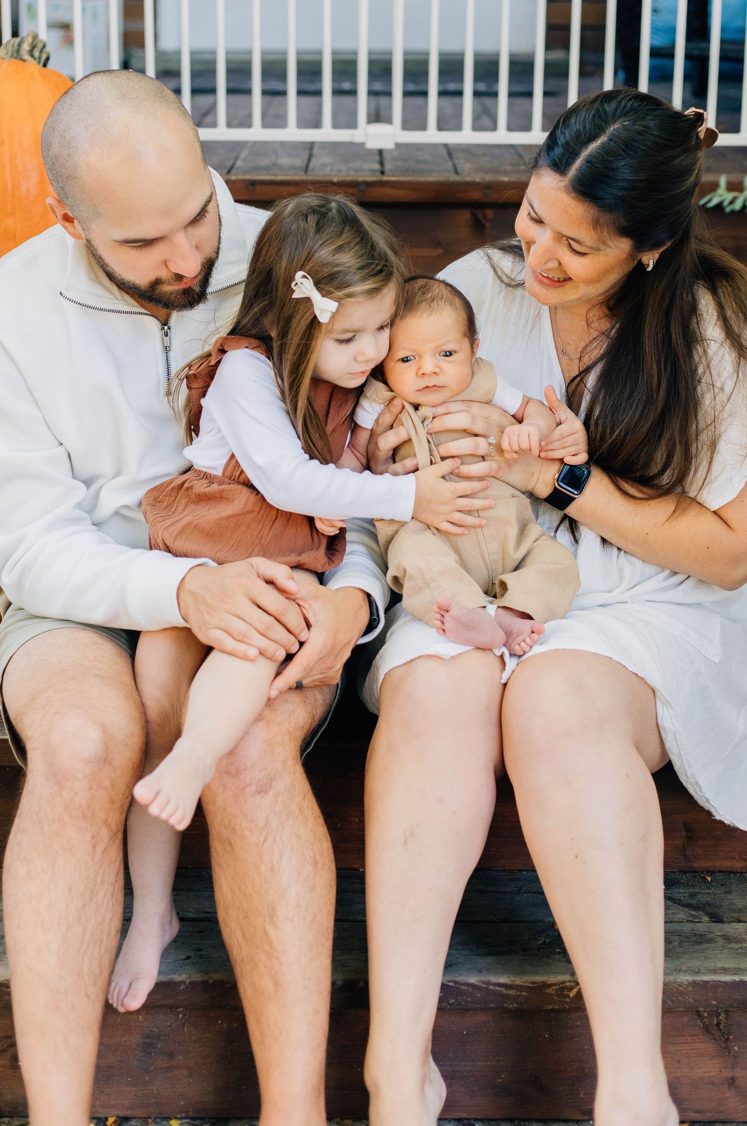 Close-up of family on wooden steps, older sister gently hugging newborn sibling while parents watch adoringly with pumpkins nearby.
