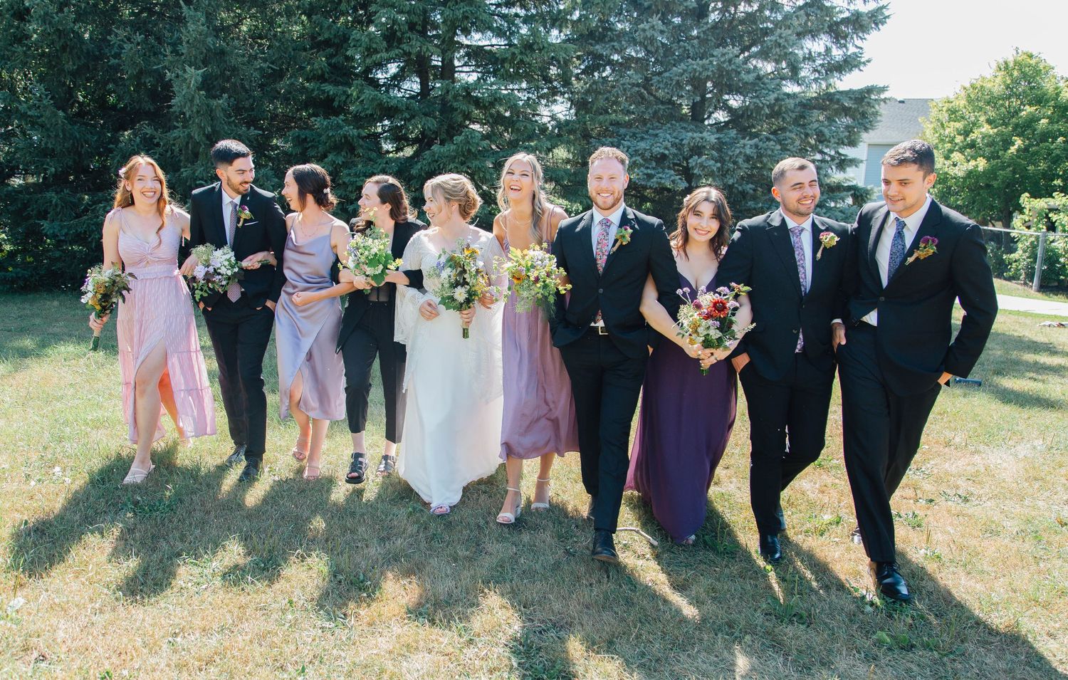 Full wedding party strolling across sunny lawn with bridesmaids in lavender and purple dresses and groomsmen in black suits.