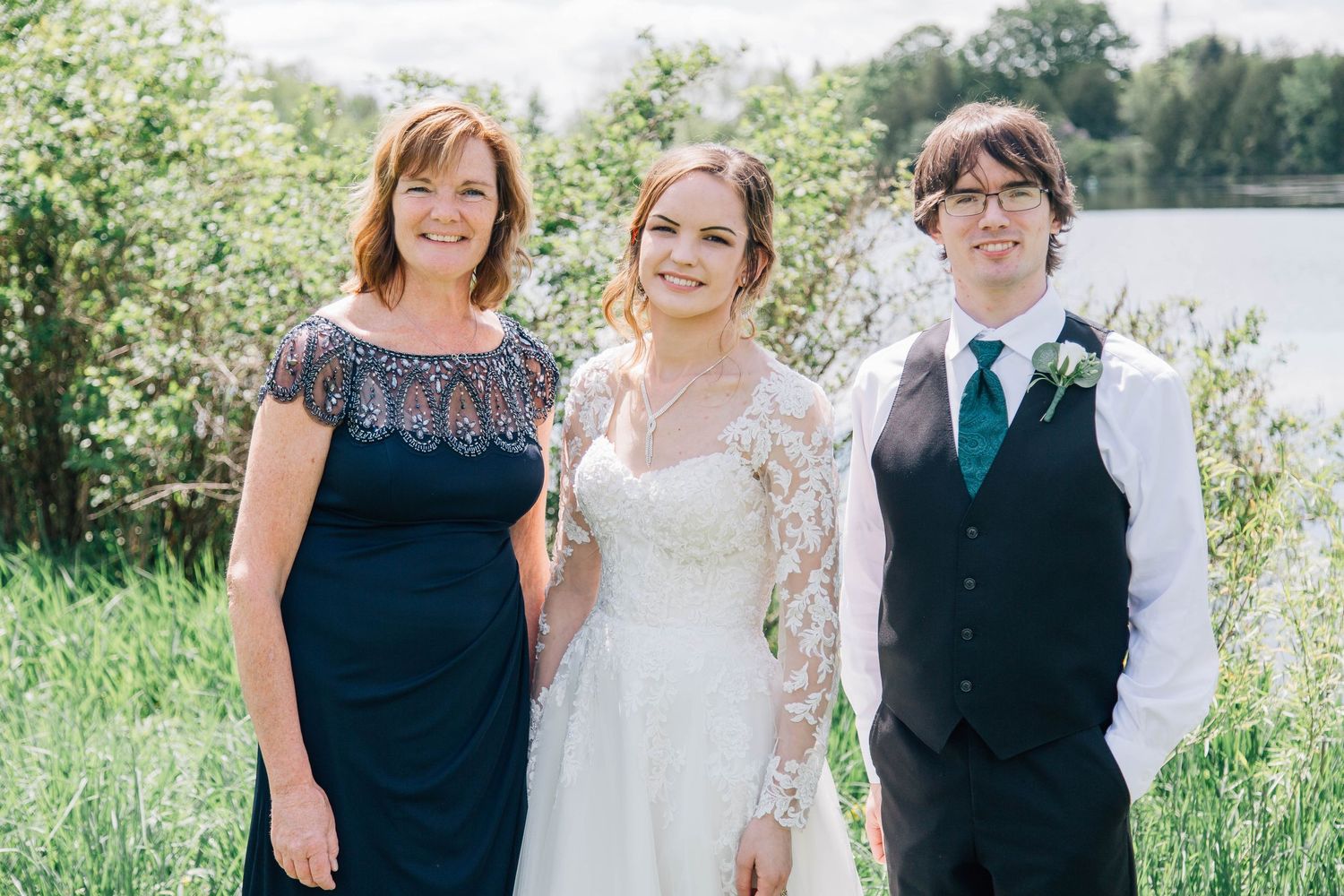 Bride in lace wedding gown posing with two family members beside a sunlit pond on wedding day.