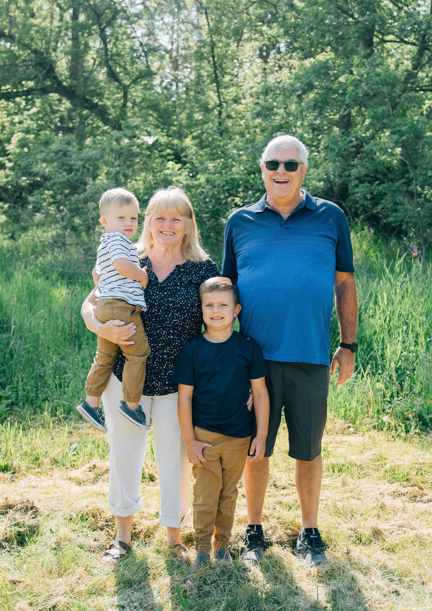 Smiling grandparents posing outdoors with two young grandsons in a lush green field on a sunny day.