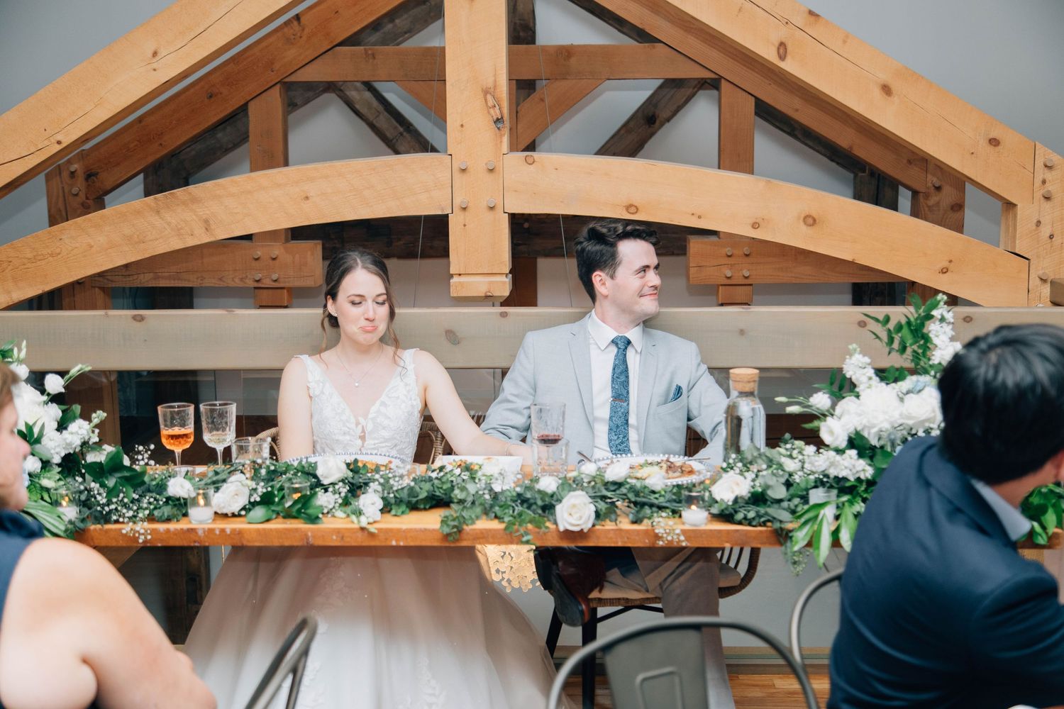Bride and groom seated at floral head table during wedding reception in rustic barn venue.
