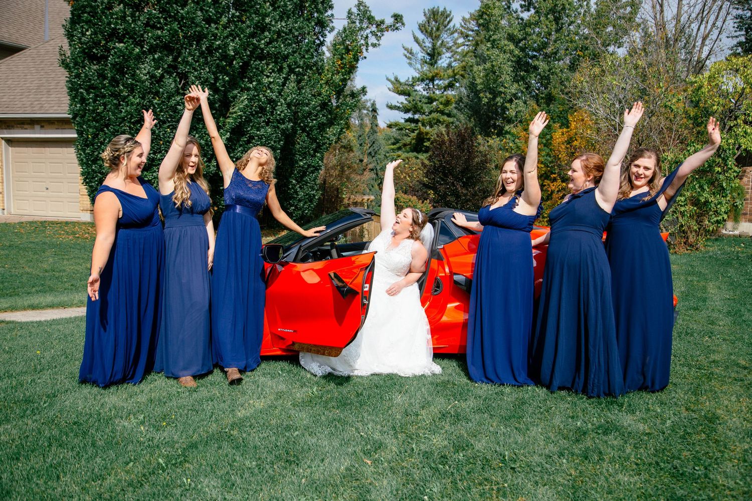 Bride and bridesmaids in royal blue gowns celebrating joyfully beside red convertible car.