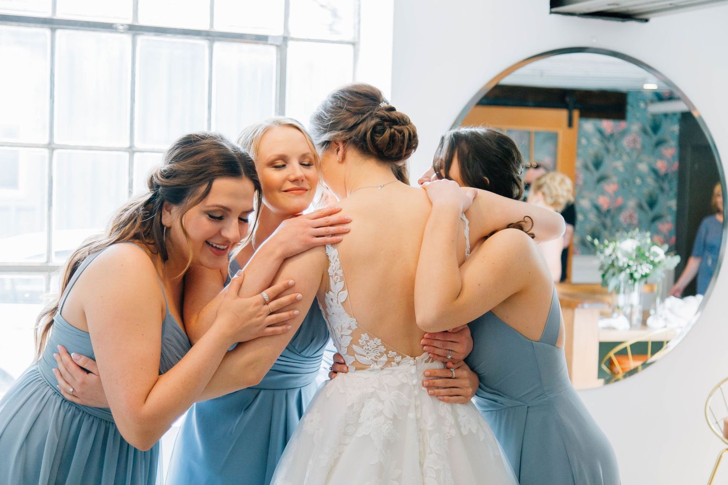 Bridesmaids in blue gowns share an emotional group hug with the bride in her lace dress.