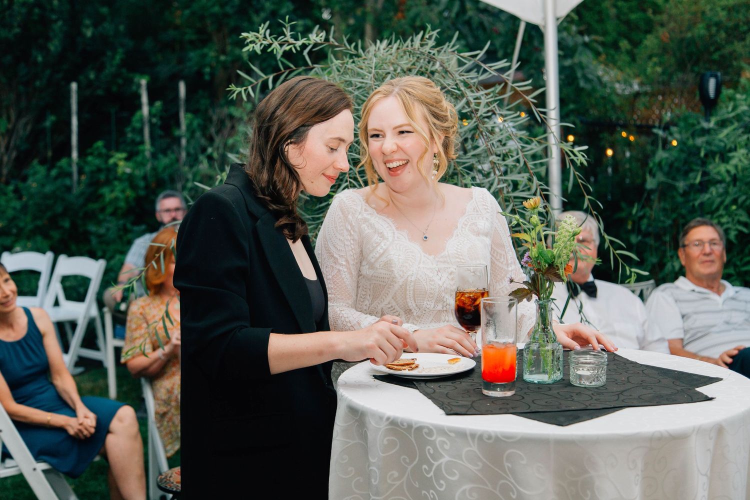 Bride in white lace dress shares a quiet moment with dark-haired guest at outdoor reception.
