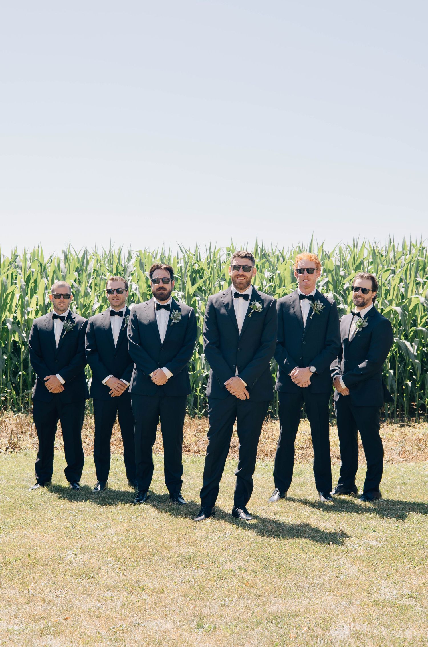 Six groomsmen wearing black tuxedos and sunglasses standing confidently in front of a green cornfield.