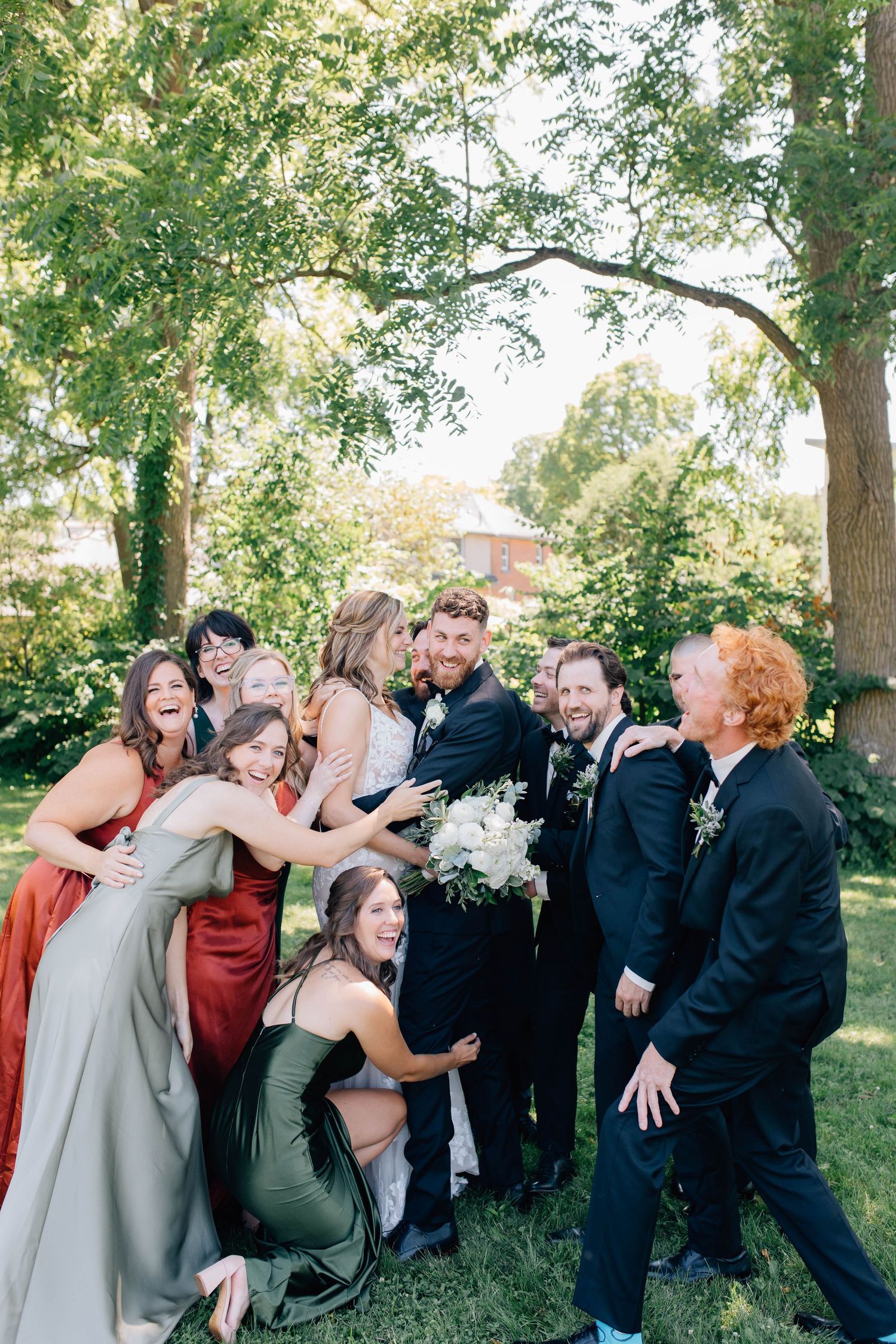 Wedding party laughing and hugging the bride and groom outdoors under green trees.