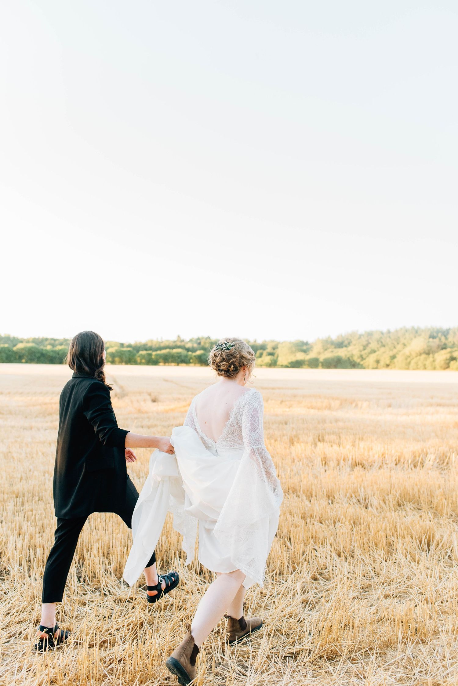 Lesbian wedding couple holding hands while walking through a golden wheat field at sunset.