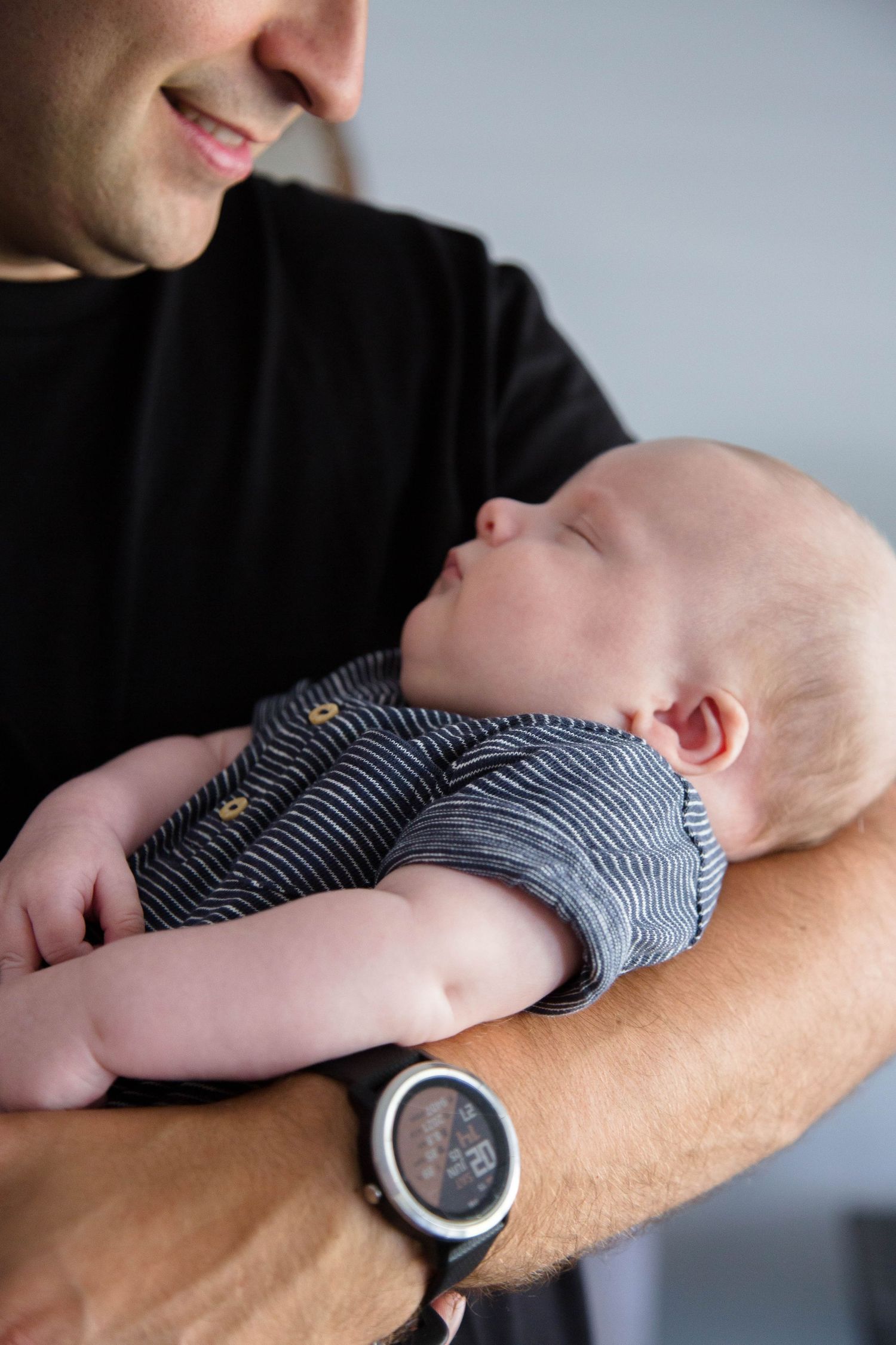 Father in black shirt holding sleeping newborn baby in striped onesie with sport watch.