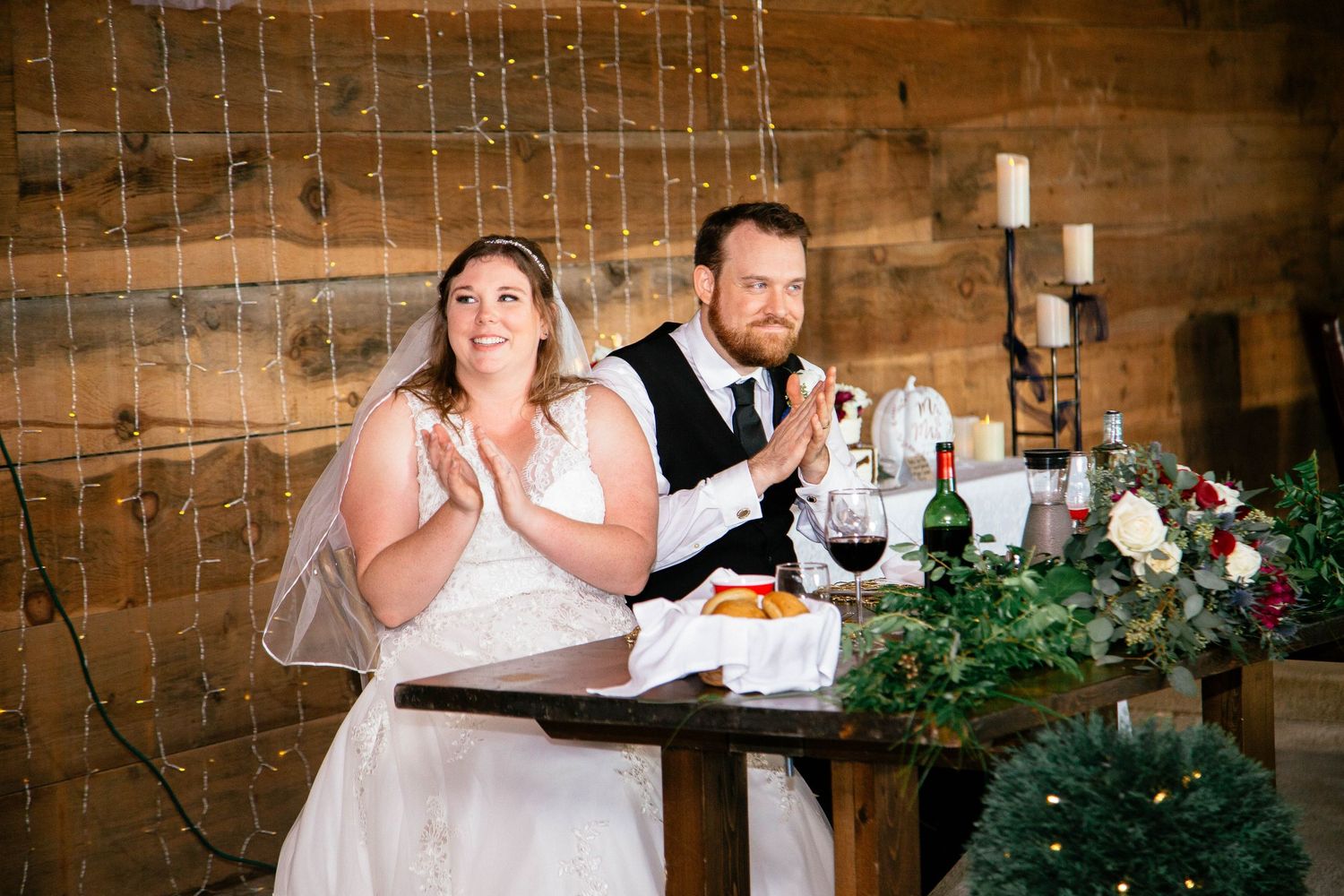 Bride and groom clapping together at rustic barn wedding reception sweetheart table with holiday greenery.
