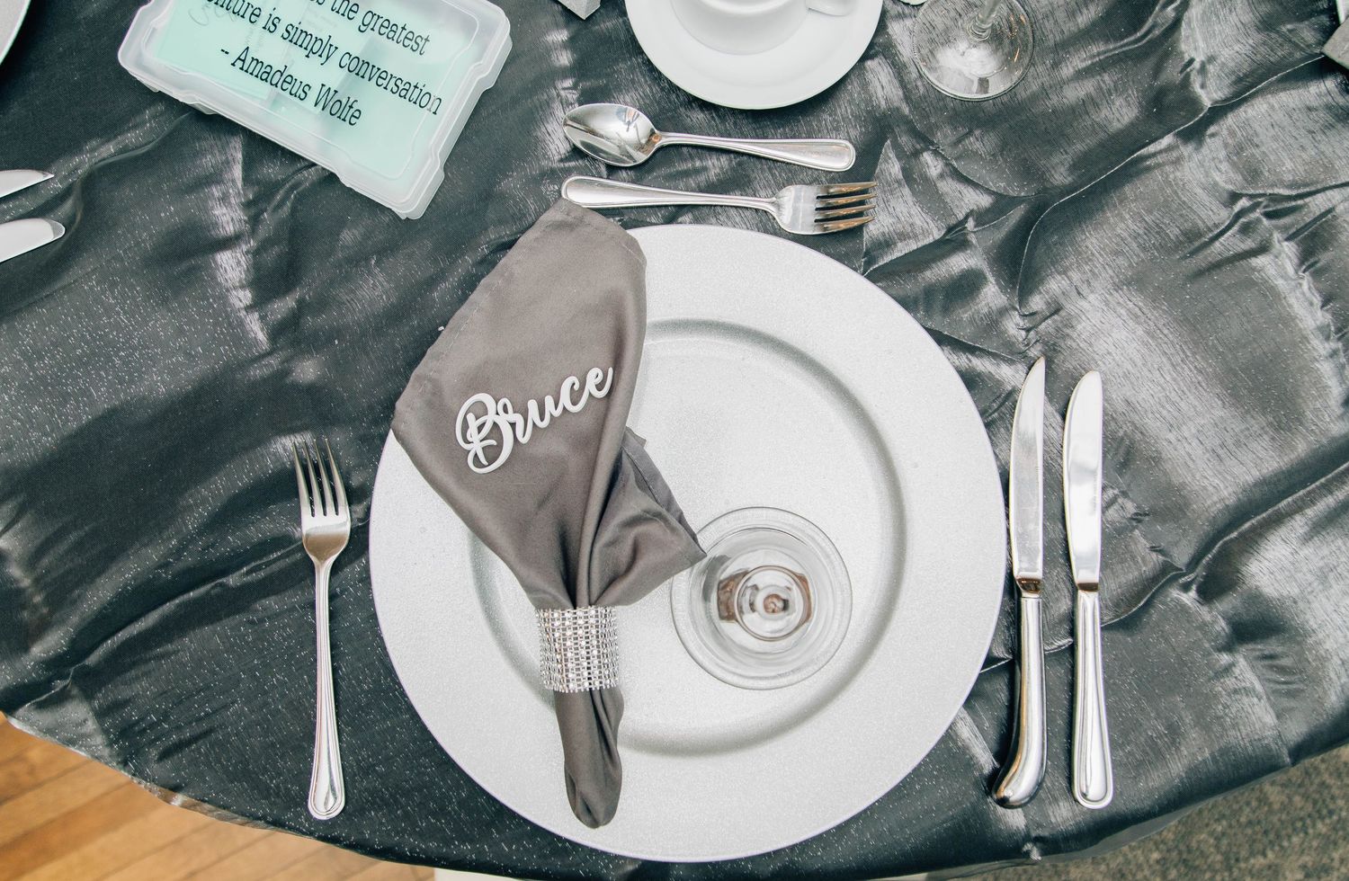 Elegant wedding place setting with embroidered Bruce napkin, white plate, and silver cutlery on gray tablecloth.