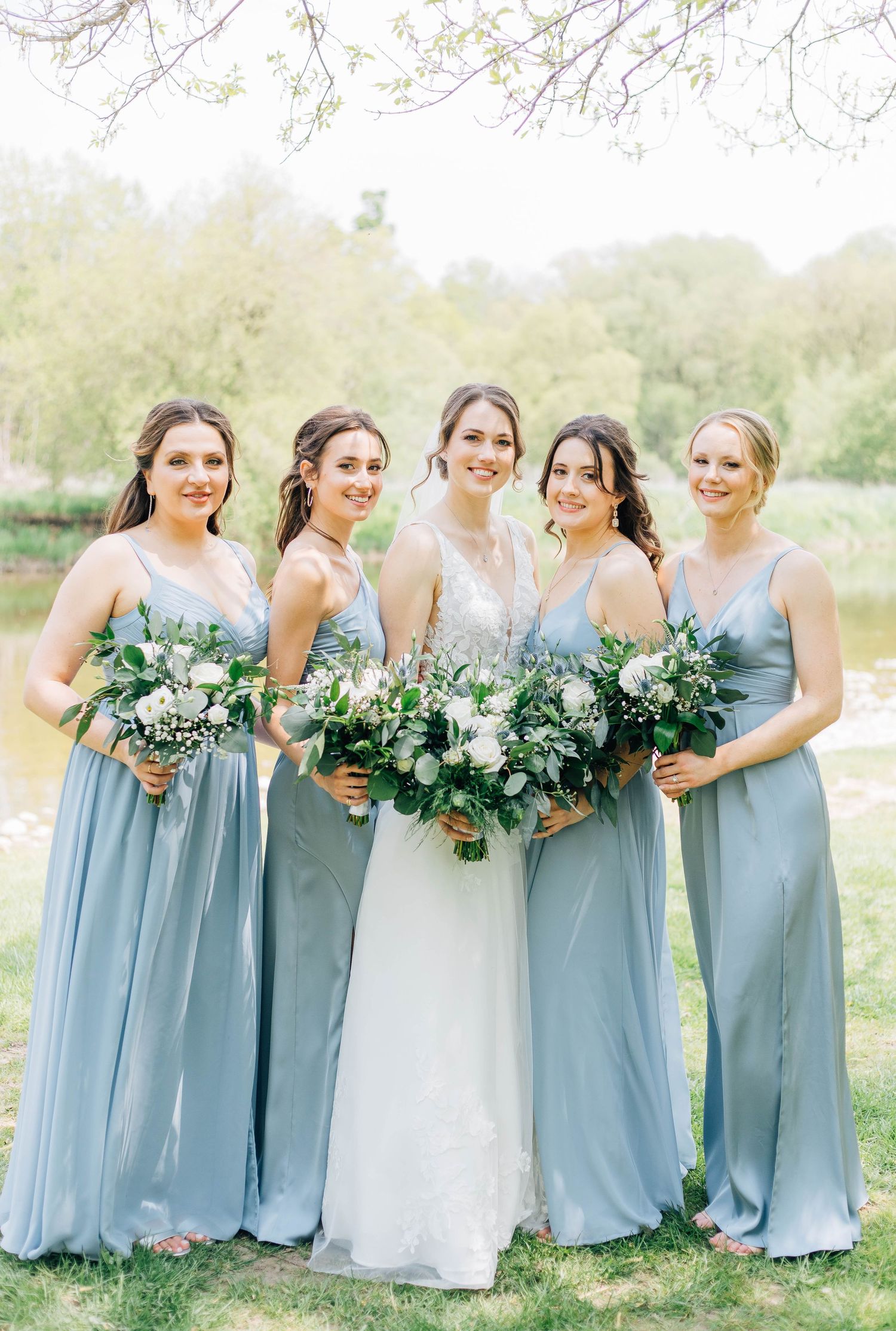 Bride in lace gown poses with four bridesmaids in dusty blue dresses holding white and green bouquets.
