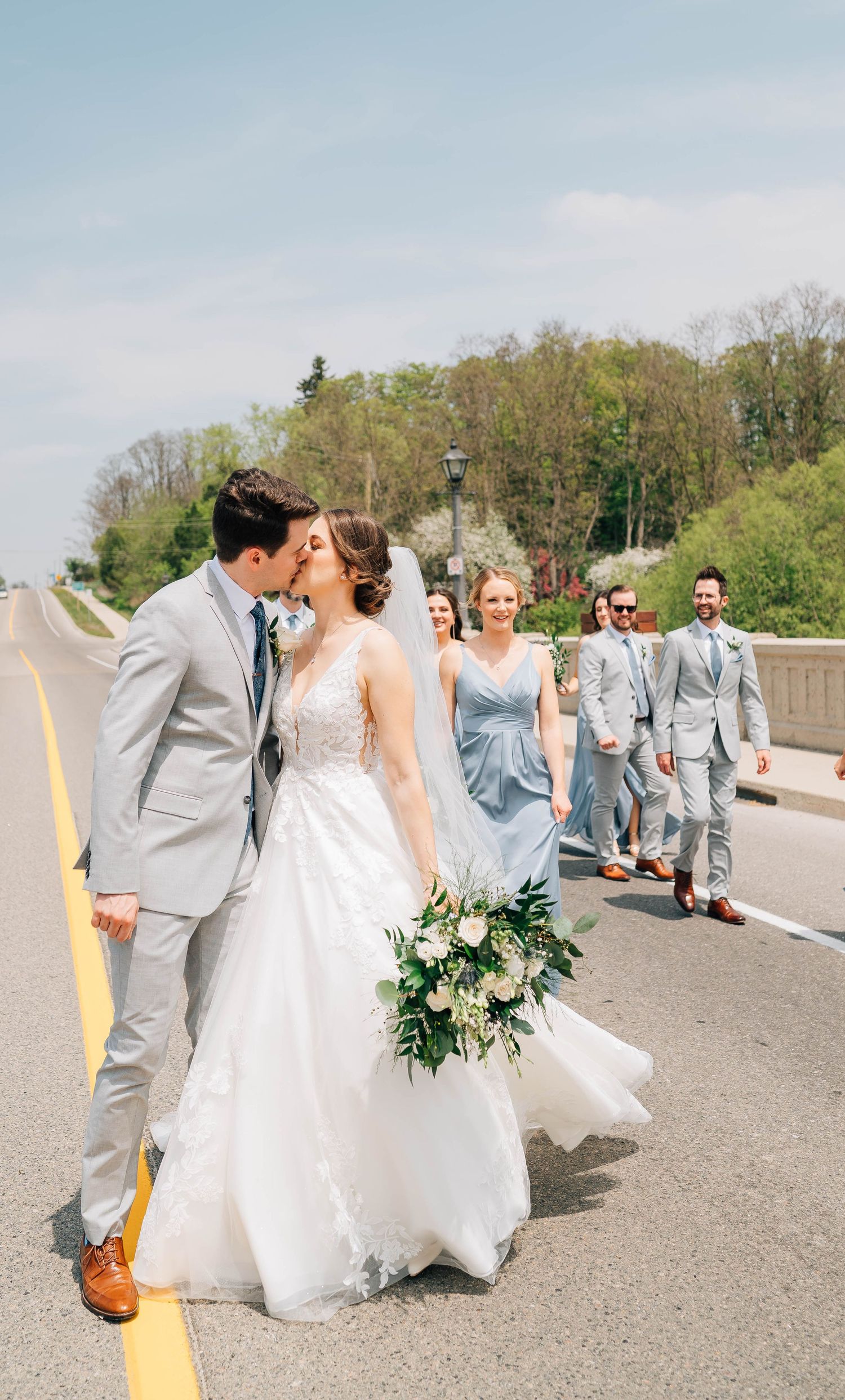 Bride and groom share a kiss on road in St. Jacob's while wedding party follows behind on sunny spring afternoon.