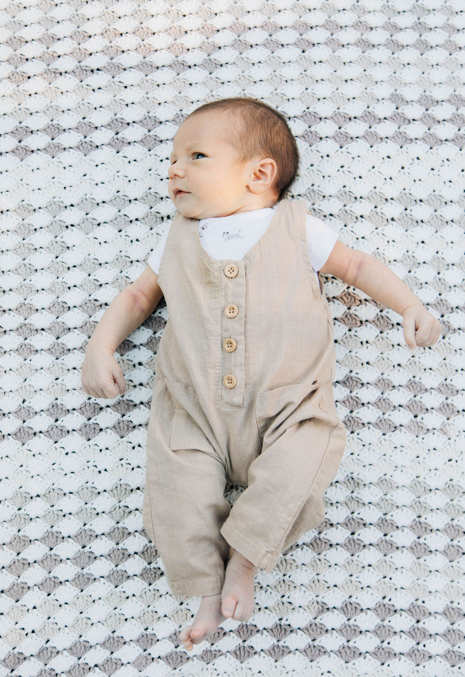 Adorable newborn baby in beige linen romper lying on white knit blanket.
