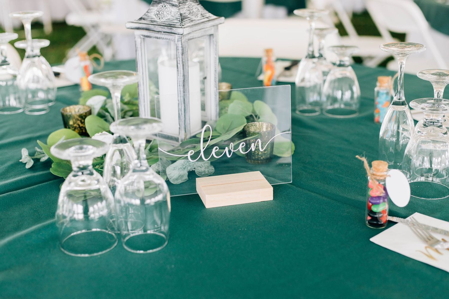 Wedding reception table eleven with green tablecloth, lantern centerpiece, and wine glasses.