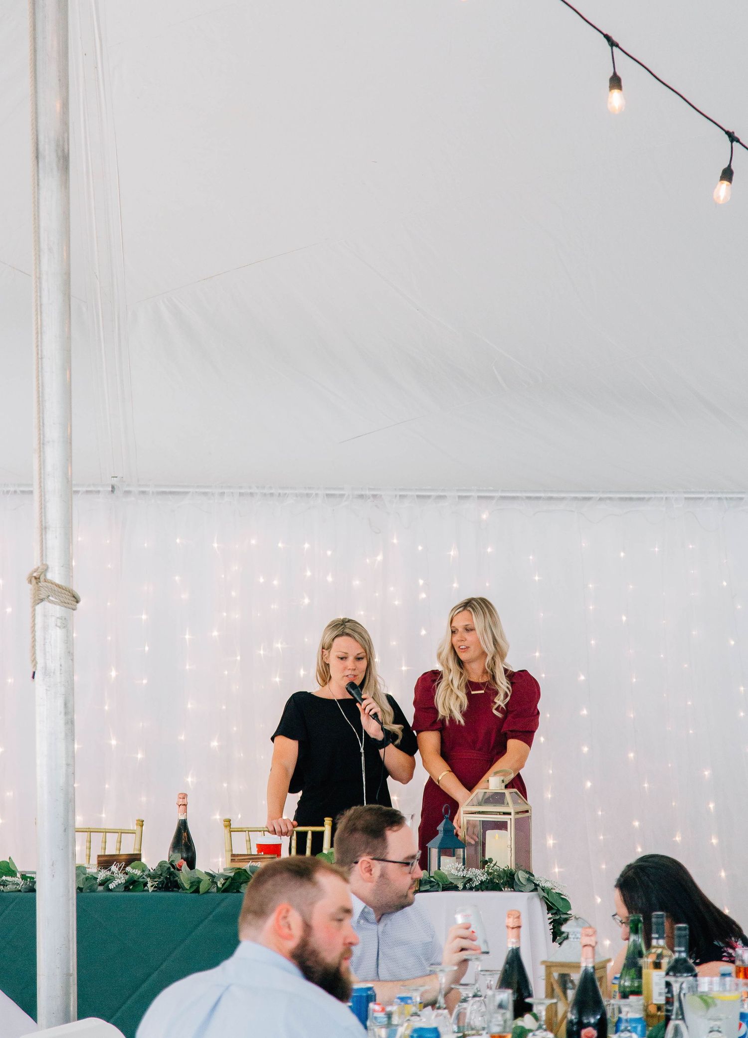 Two women share a microphone while giving a toast at a wedding reception under string lights and a white tent.